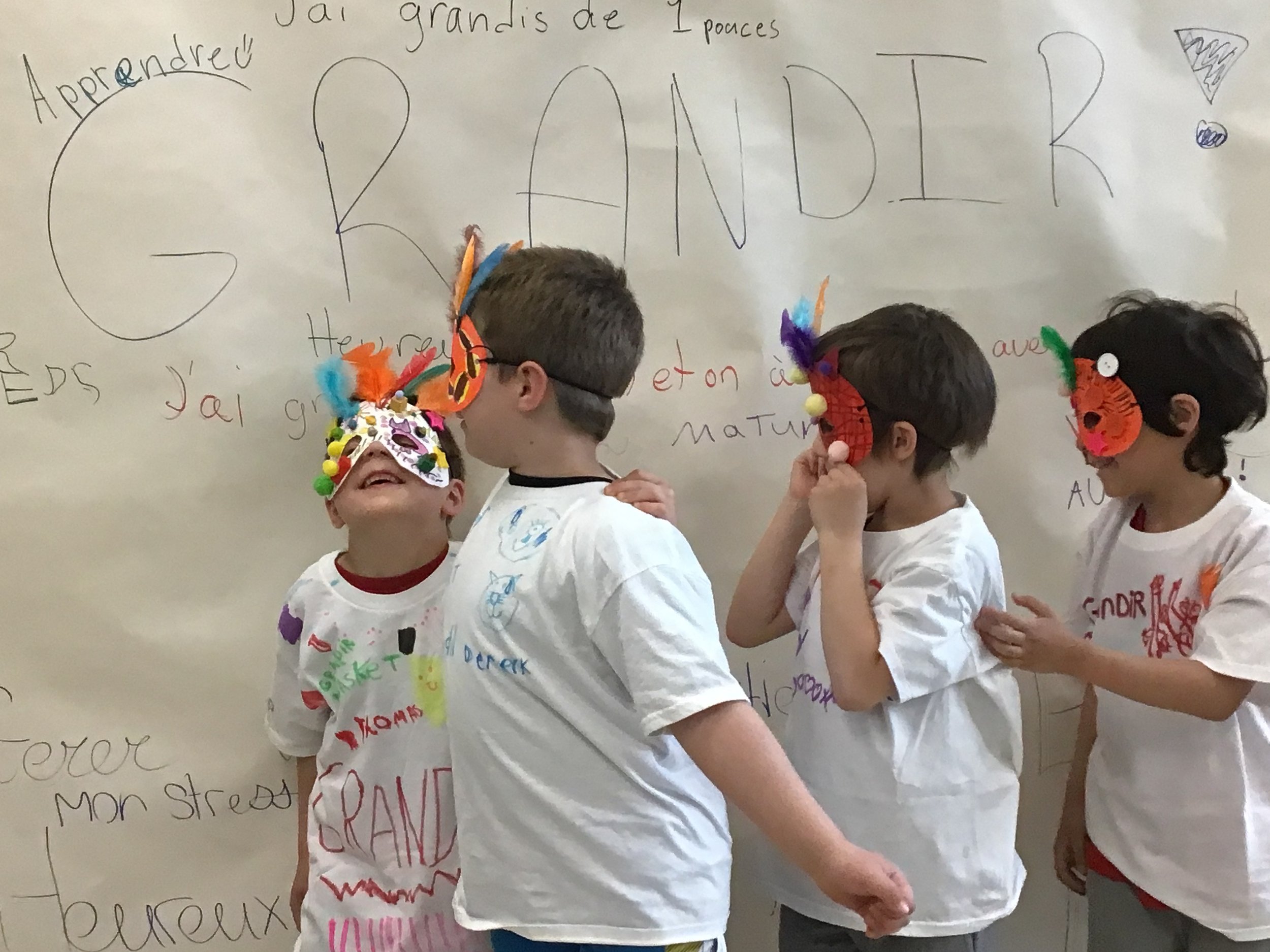 Quatre enfants portant des masques colorés et des t-shirts blancs écrits à la main, se déroulant devant un mur avec des écrits en français, participant à une activité artistique ou théâtrale.