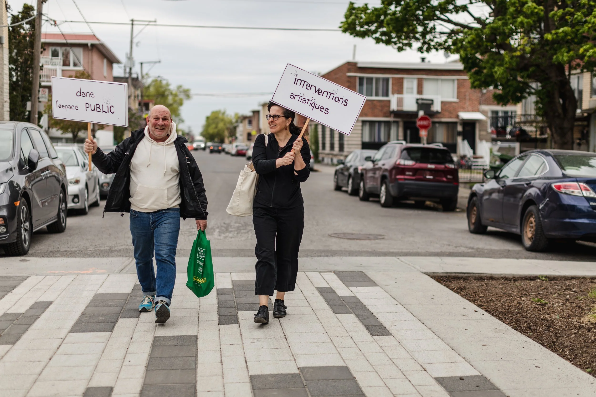 Deux personnes marchent dans la rue avec des pancartes en soutien à l'intervention artistique dans l'espace public, sous un ciel nuageux, avec des voitures stationnées sur le côté et des bâtiments résidentiels en arrière-plan.