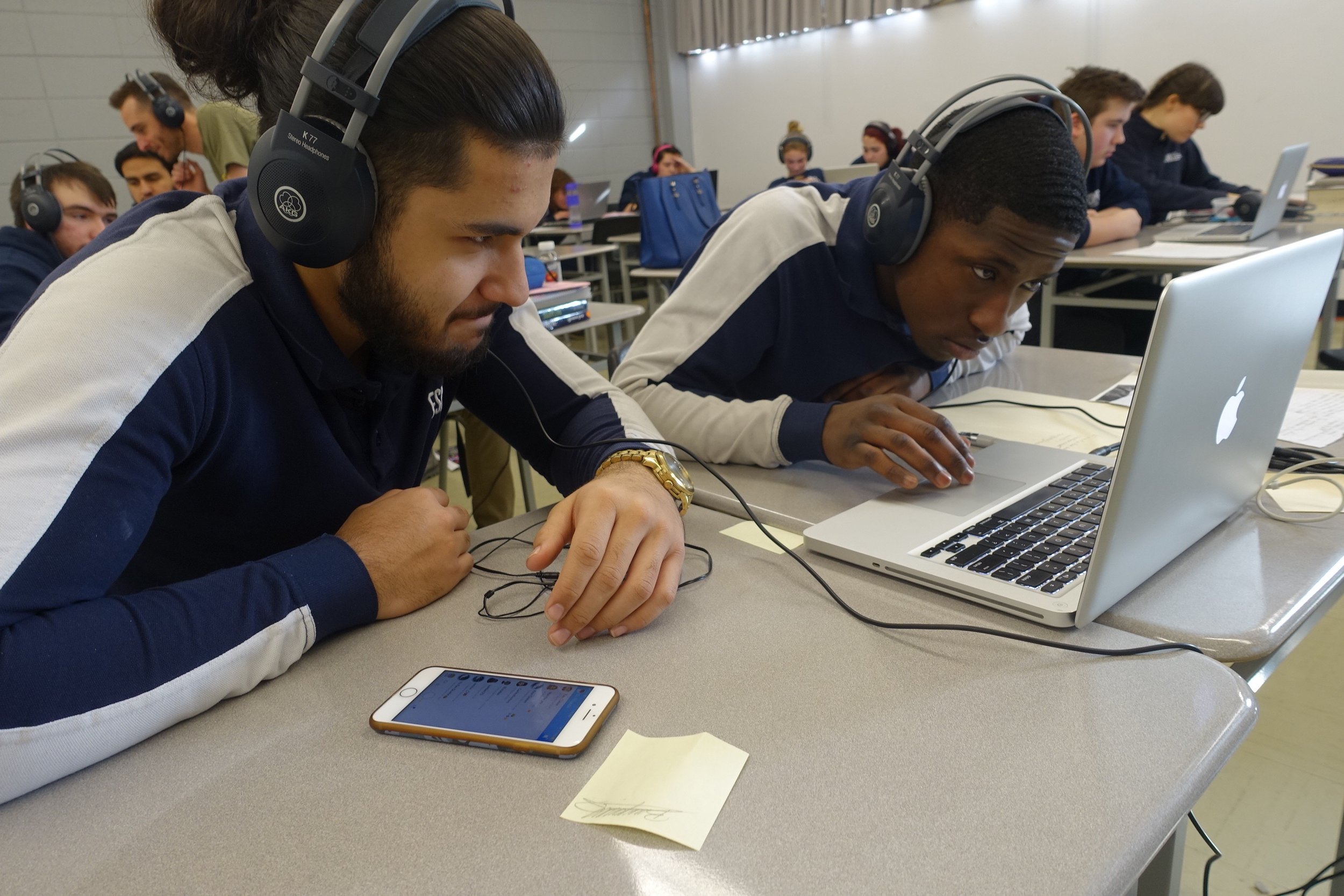 Deux jeunes hommes portant des écouteurs concentrés devant un ordinateur portable dans une salle de classe ou une bibliothèque avec d'autres étudiants en arrière-plan.