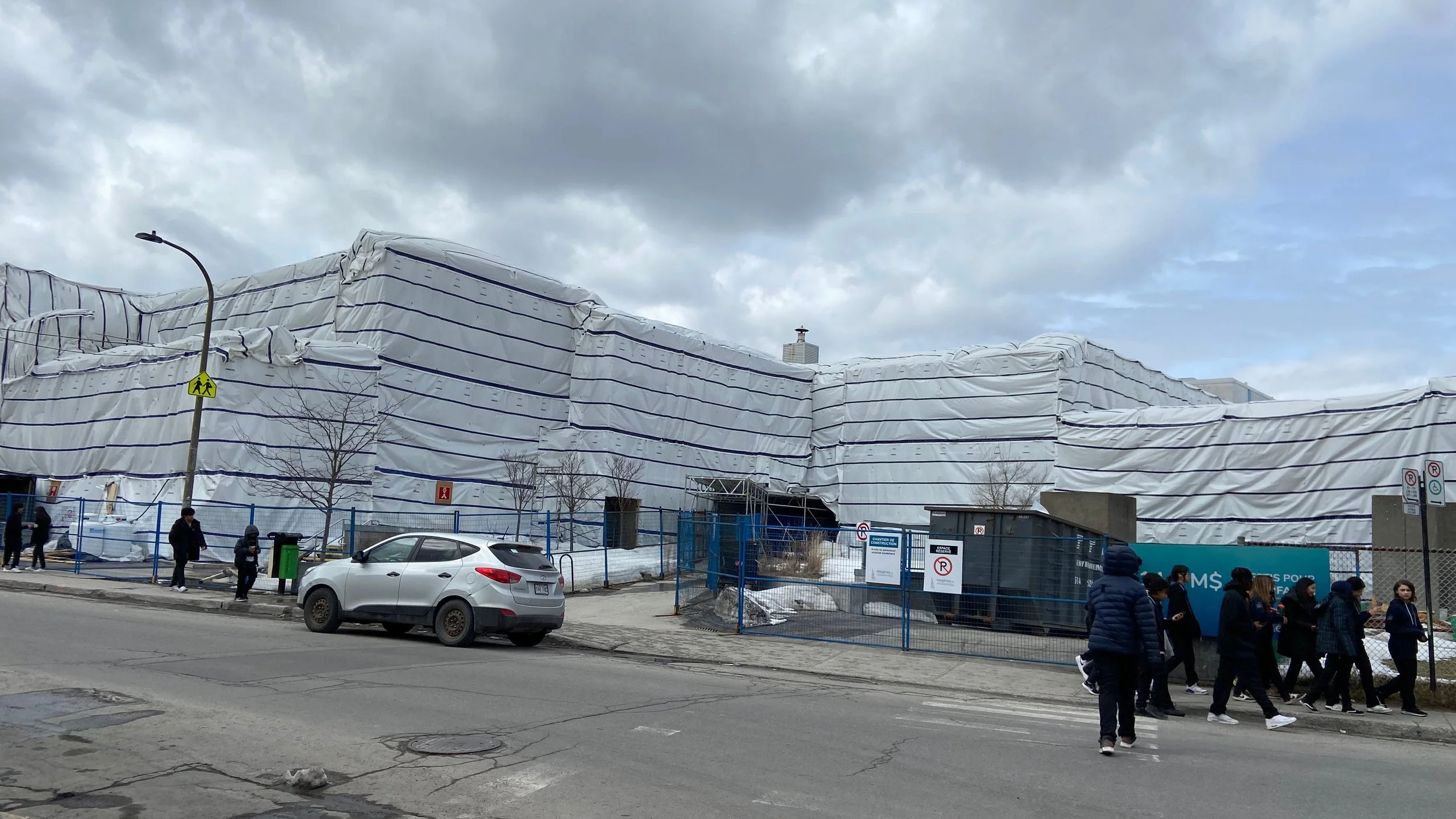 Construction en cours avec un bâtiment entouré d'une grande bâche blanche et des piétons marchant sur le trottoir en face.