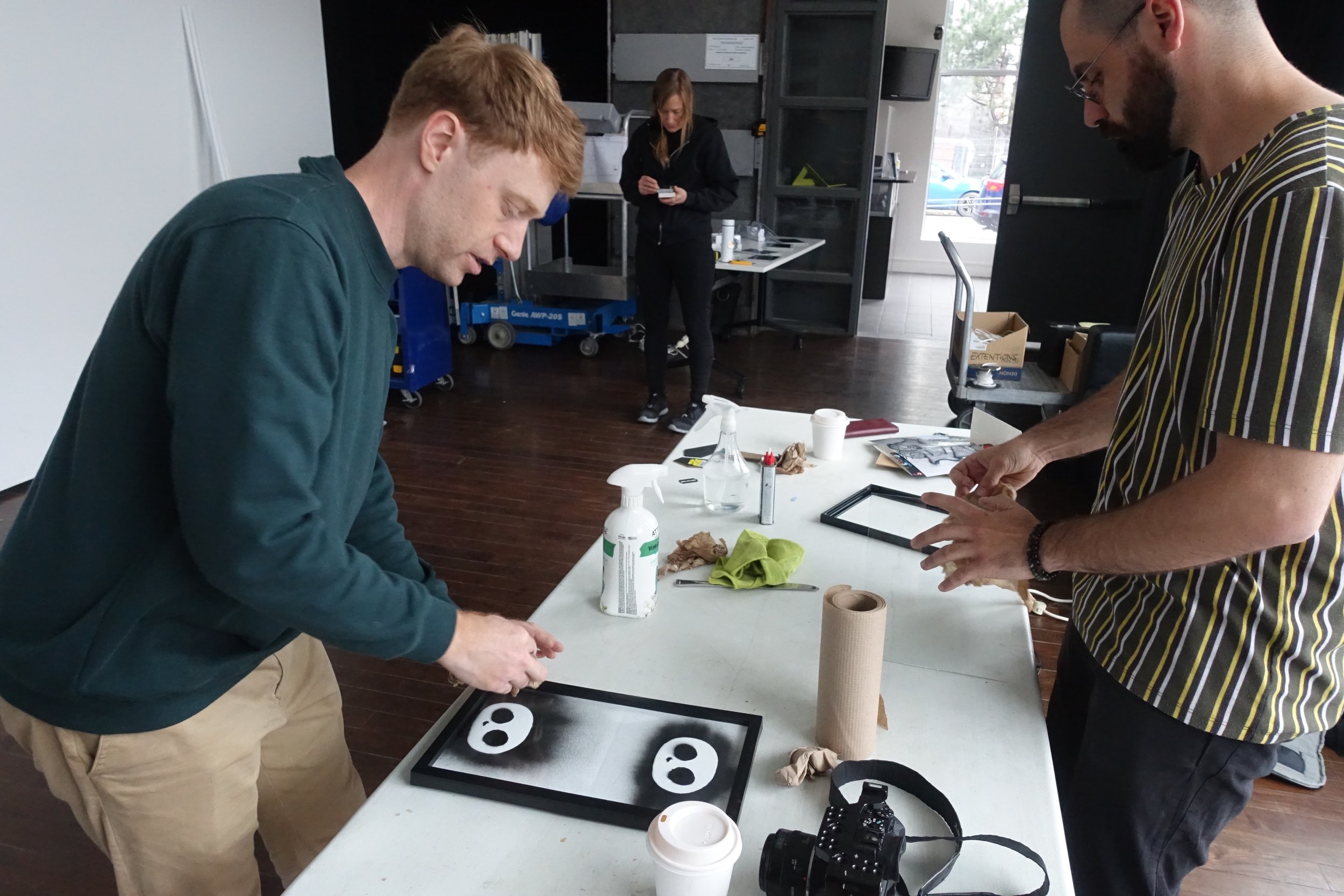 Deux hommes travaillent sur une table avec des masques en carton, des produits de nettoyage et un appareil photo. Une femme en arrière-plan regarde son téléphone dans un espace industriel.