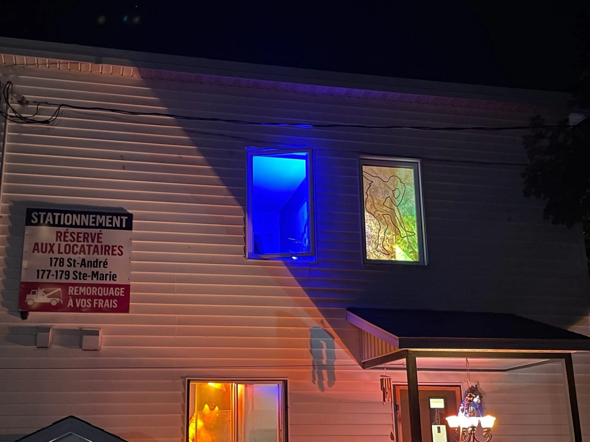 Façade d'un bâtiment résidentiel avec deux fenêtres, une illuminée en bleu et une autre en jaune, une enseigne de stationnement réservée aux locataires et un petit porche avec une lampe suspendue devant la porte d'entrée.
