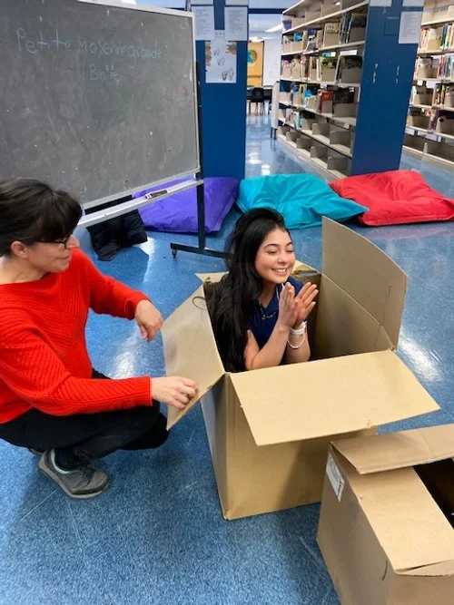 Deux jeunes filles jouent dans une grande boîte en carton dans une bibliothèque, l'une avec un sweat rouge et l'autre avec une chemise bleue, toutes deux souriantes.