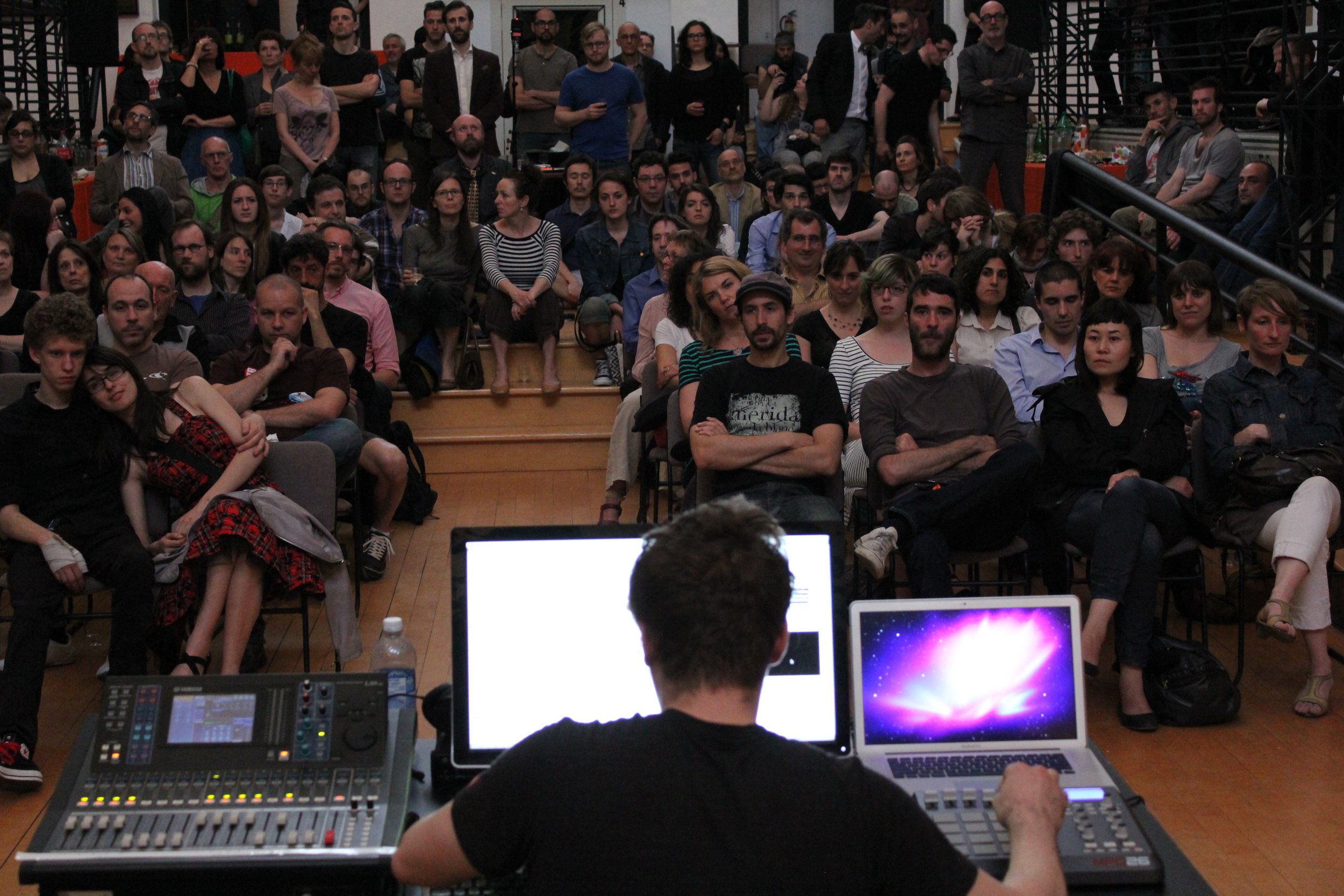 Une personne dirige une présentation ou un événement devant une large audience dans une salle de conférence ou un auditorium. L'arrière-plan montre un public assis, attentif. Plusieurs ordinateurs et équipements électroniques sont visibles sur la tab