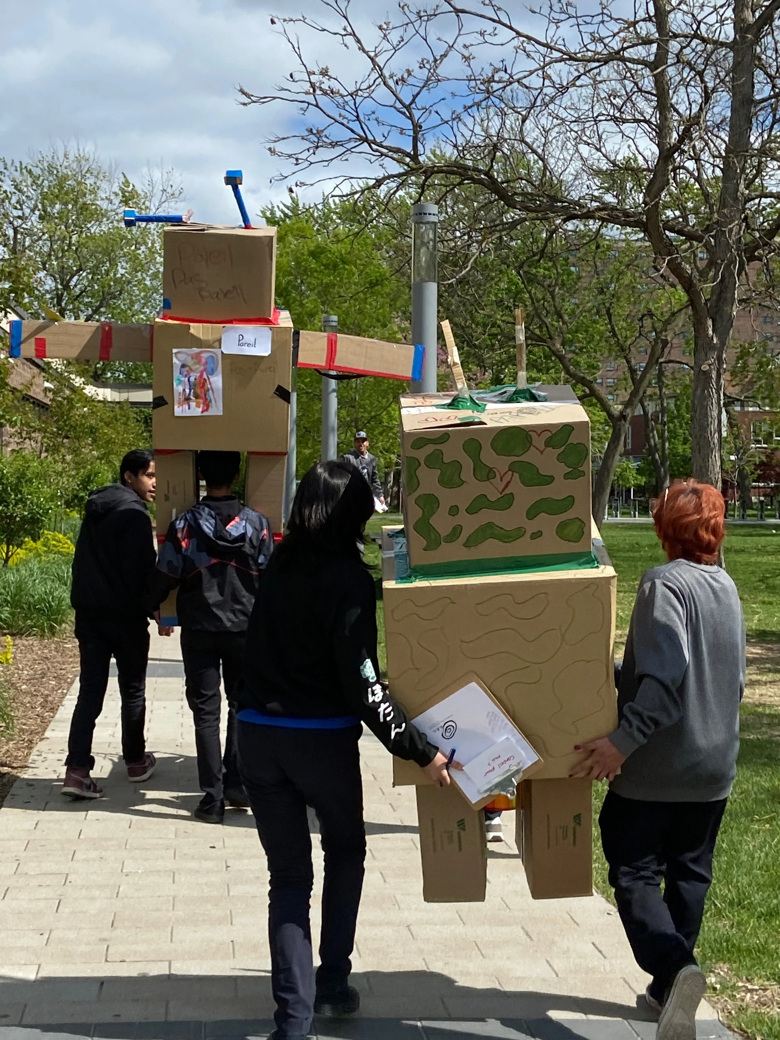 Groupe de personnes transportant une grande sculpture en carton dans un parc. La sculpture ressemble à un avion ou un hélicoptère en utilisant diverses boîtes en carton.
