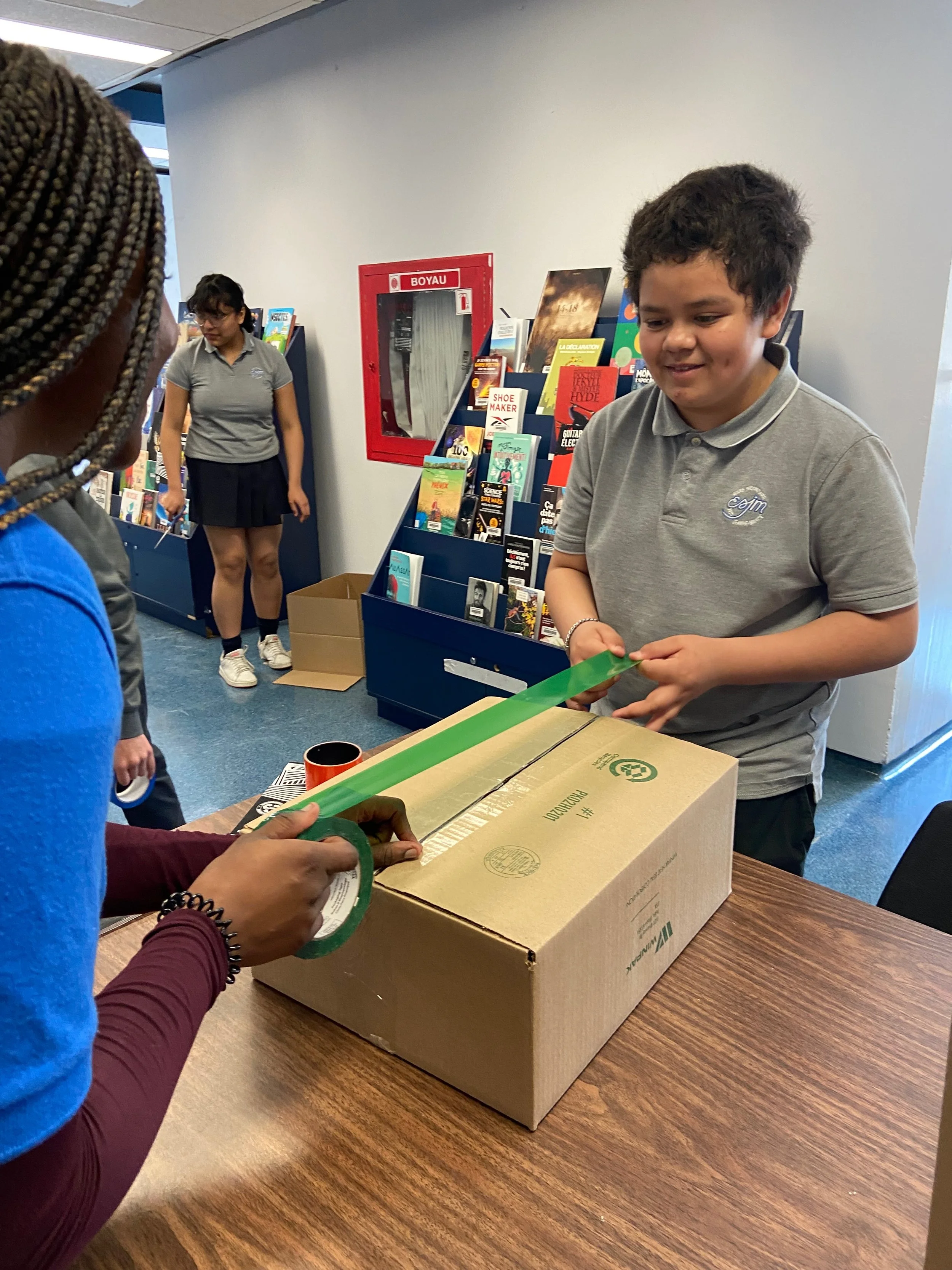 Une personne utilisant du ruban adhésif pour fermer un colis en carton, avec deux enfants qui regardent, dans un environnement intérieur.
