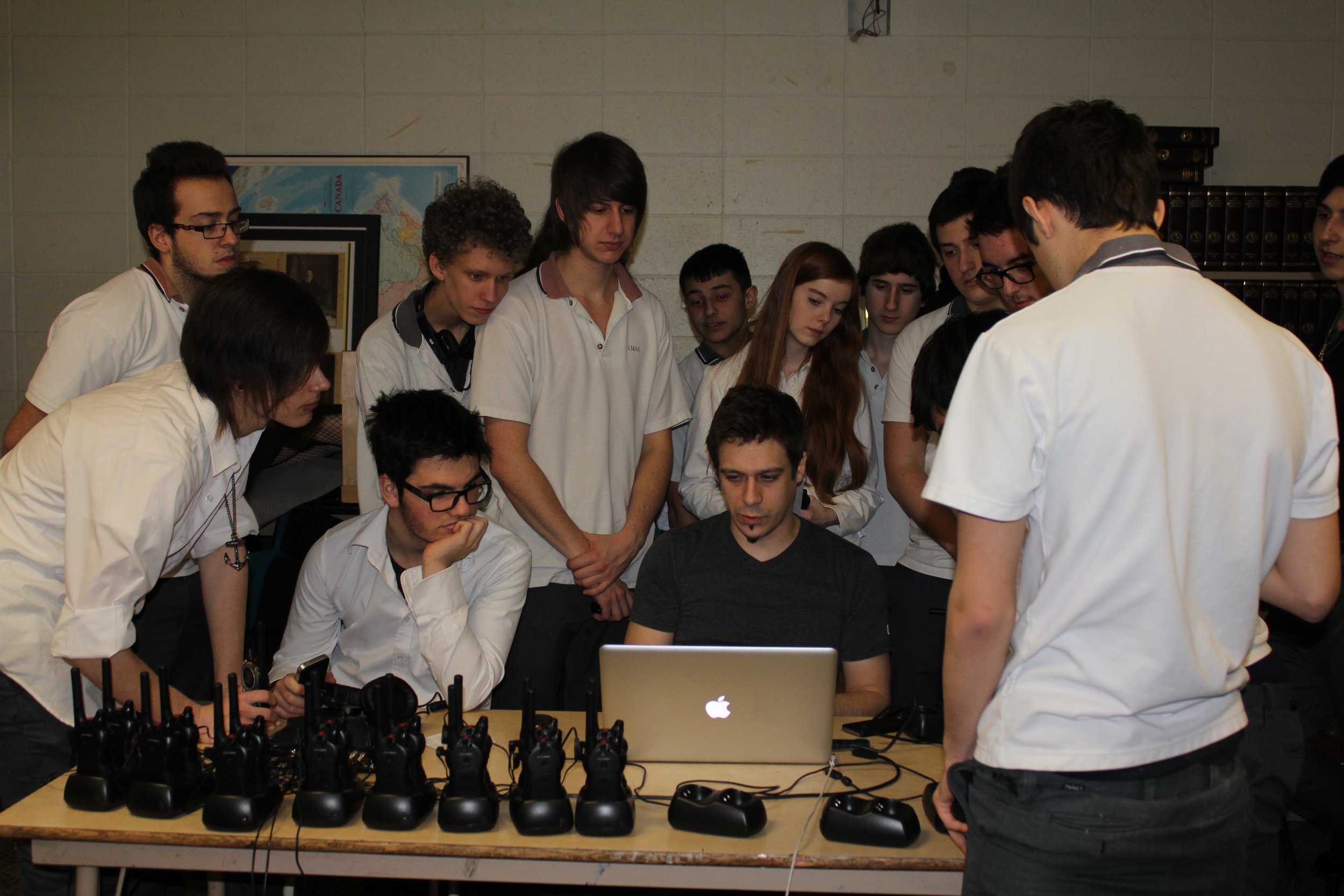 Groupe de jeunes étudiants autour d'un ordinateur portable, en train d'écouter une personne qui leur explique quelque chose, avec plusieurs talkies-walkies sur la table.