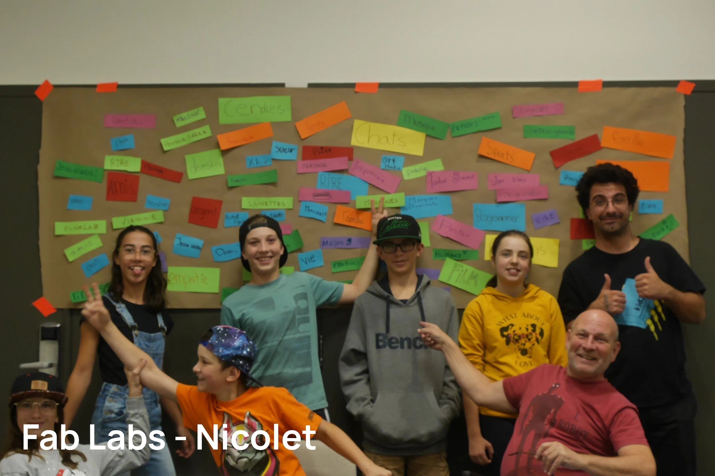 Groupe de huit personnes posant devant un tableau coloré avec des notes adhésives. Certains font des gestes de victoire ou de paix, tous souriants.