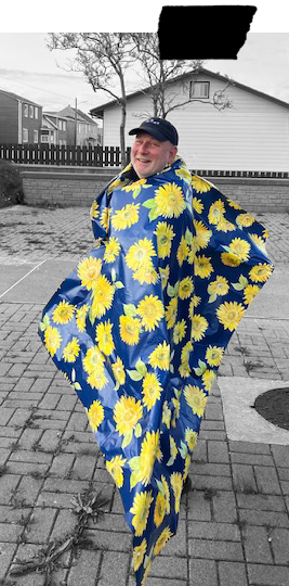 Yves Amyot souriant portant une tente à motifs de marguerites jaunes et bleues, debout dans une cour pavée en extérieur.