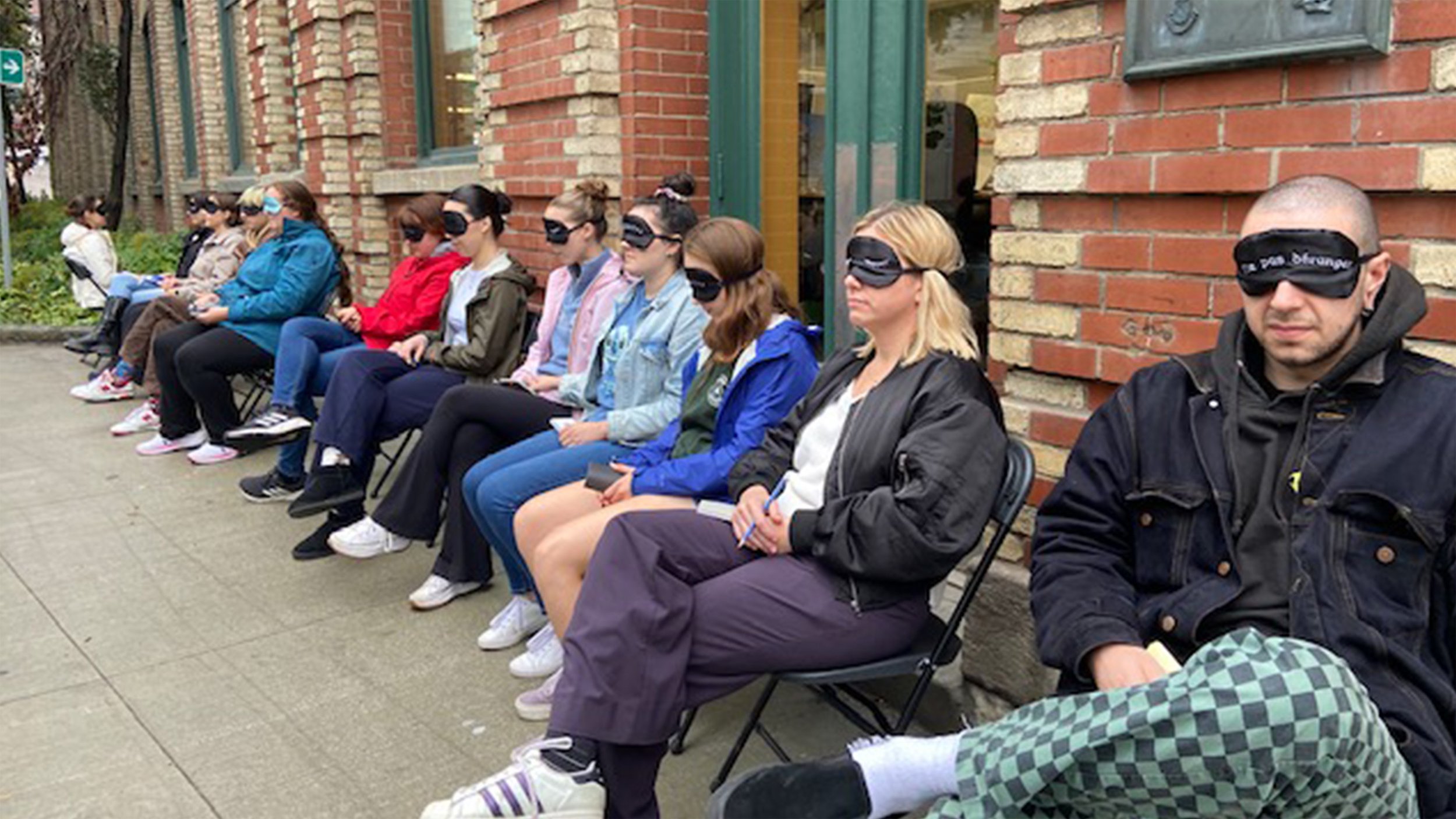 Groupe de personnes assises sur des chaises devant un mur en brique, portant des masques pour les yeux dans un contexte d'activités de relaxation ou de méditation en extérieur.