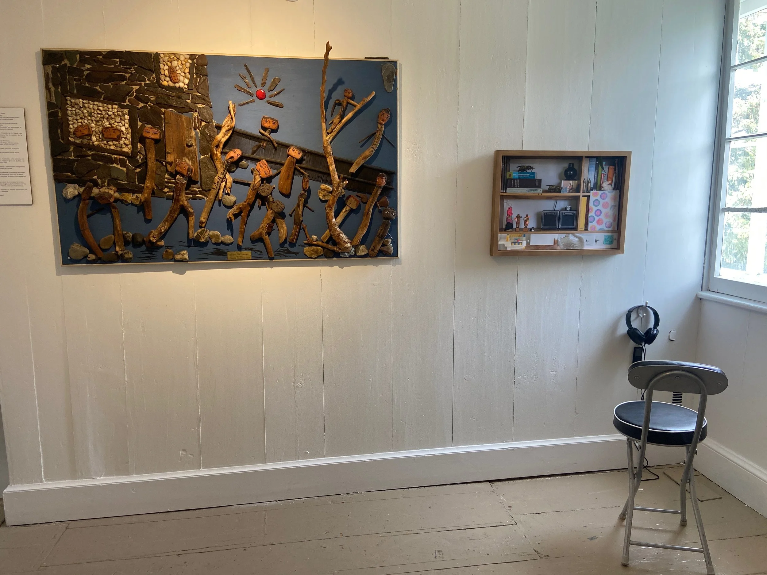 Une pièce avec une œuvre d'art murale en bois et cailloux, une étagère avec des objets, un casque audio suspendu à une chaise, et une grande fenêtre laissant entrer la lumière naturelle.