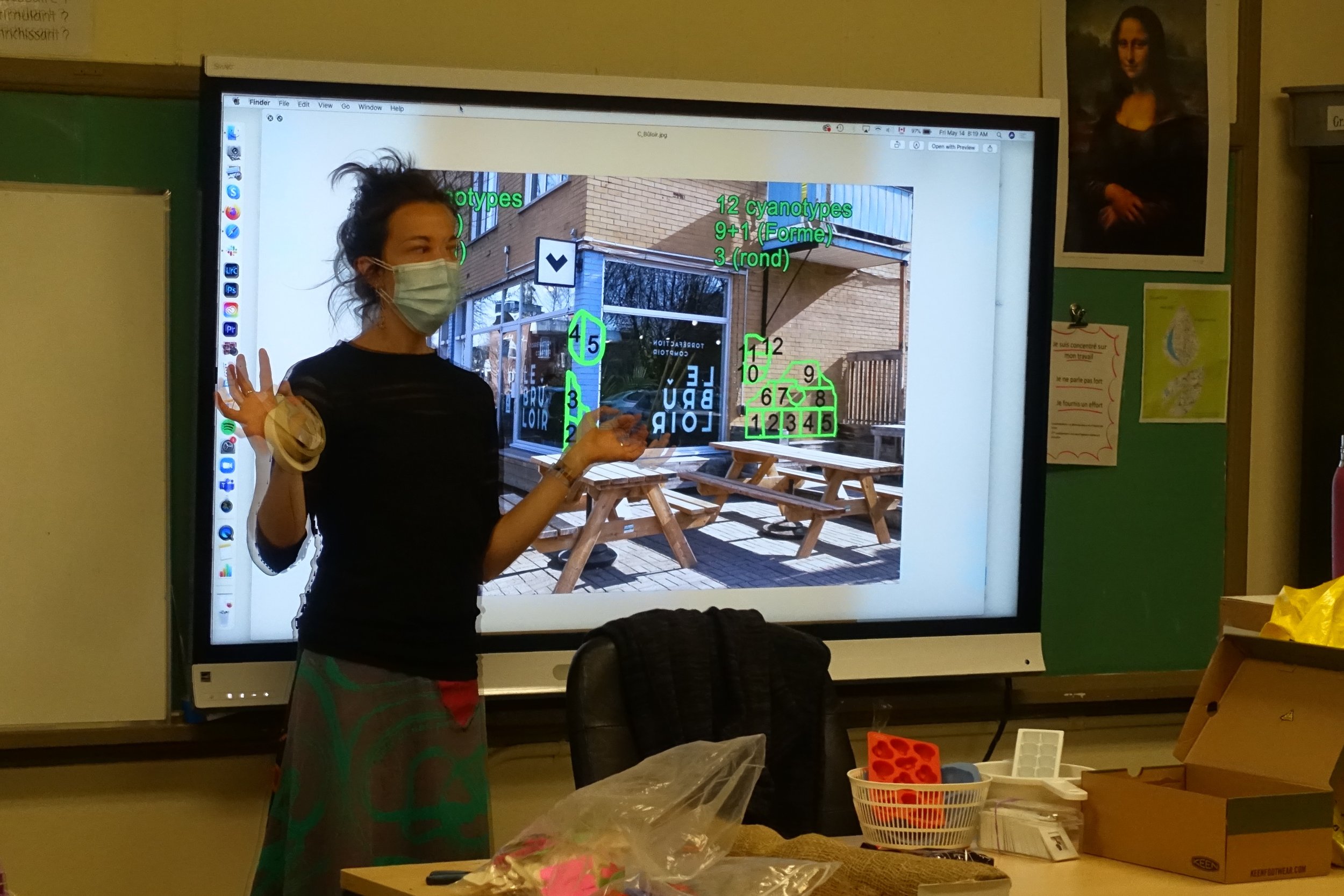Une femme portant un masque facial se tenant devant un tableau blanc interactif dans une salle de classe, avec des lunettes et une jupe colorée, en train d'expliquer quelque chose.