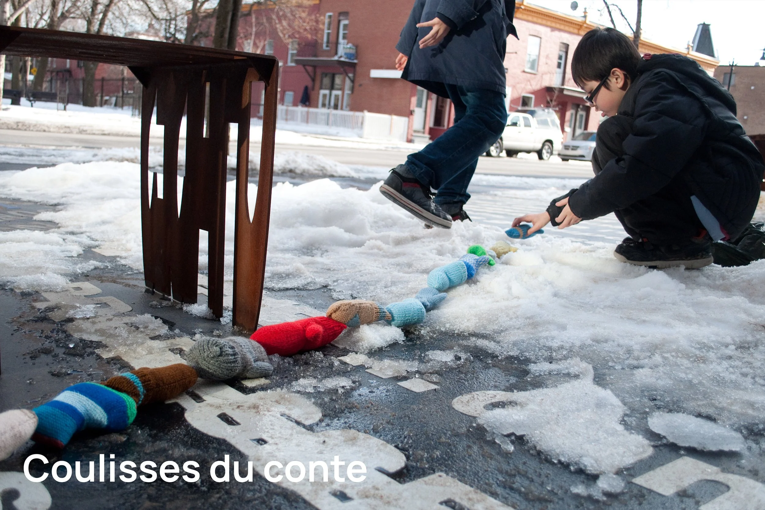 Deux enfants jouent dans la neige sur un trottoir, suspendant des chaussettes colorées à une corde tendue entre un meuble et un étage. La scène se déroule dans une zone résidentielle urbaine en hiver.