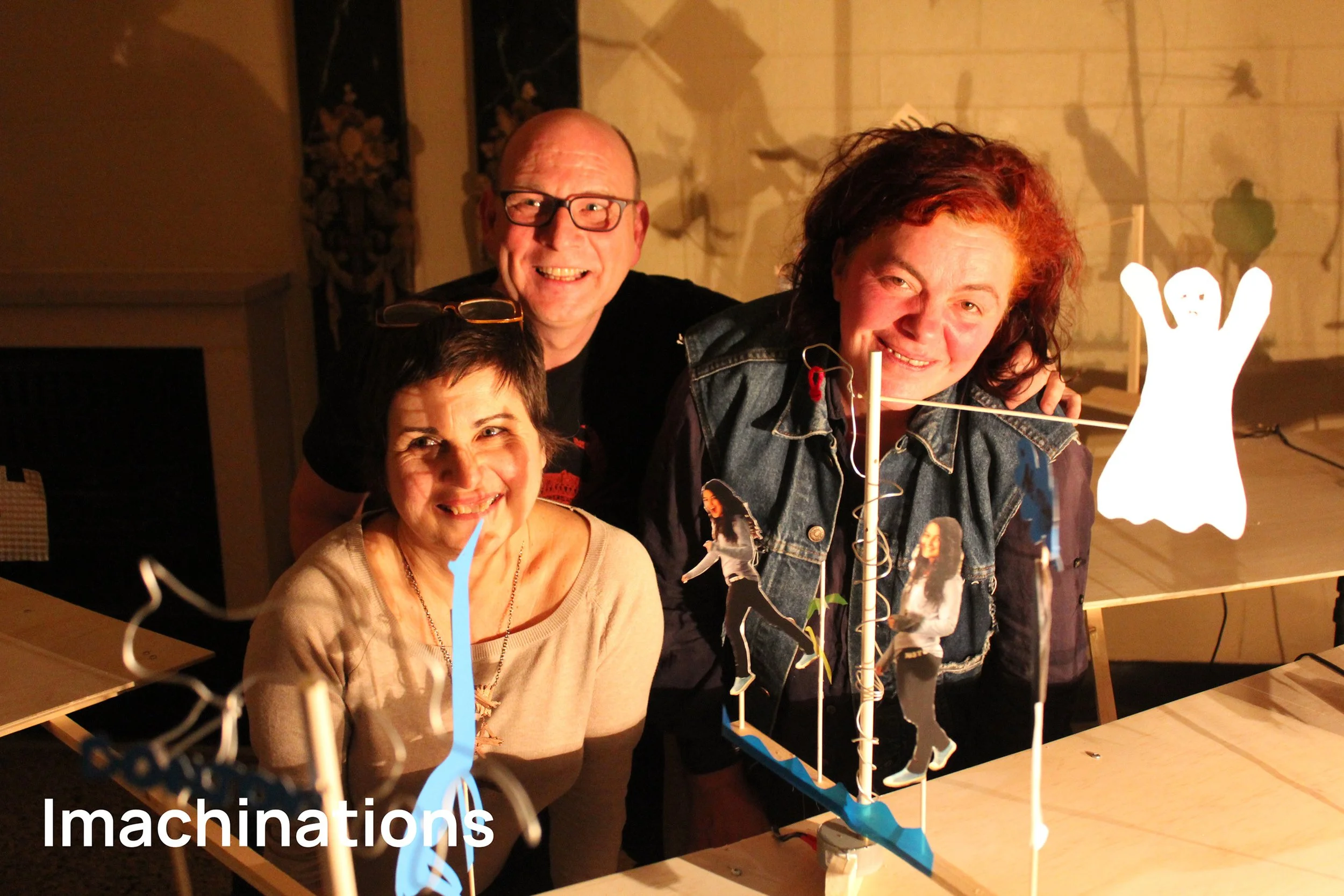 Trois personnes souriantes posant devant une création artistique en papier représentant des personnages en papier et une silhouette de fantôme, dans une pièce avec un éclairage chaud.
