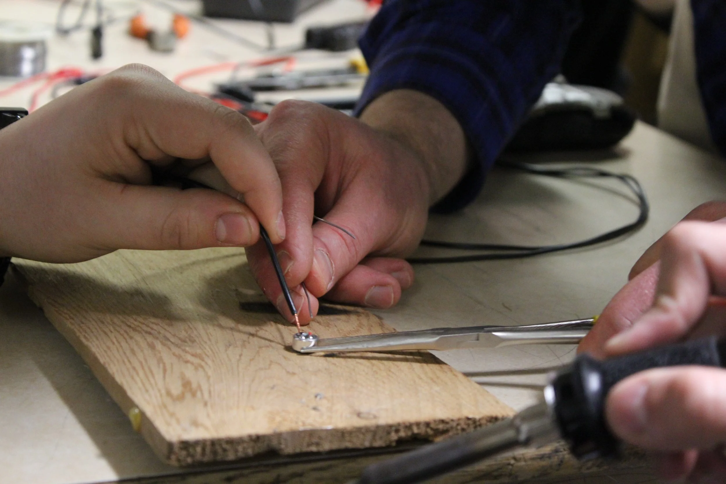 Deux personnes travaillent ensemble à l'assemblage ou la réparation d'un petit objet électronique ou mécanique sur une planche de bois, en utilisant des outils et des fils.