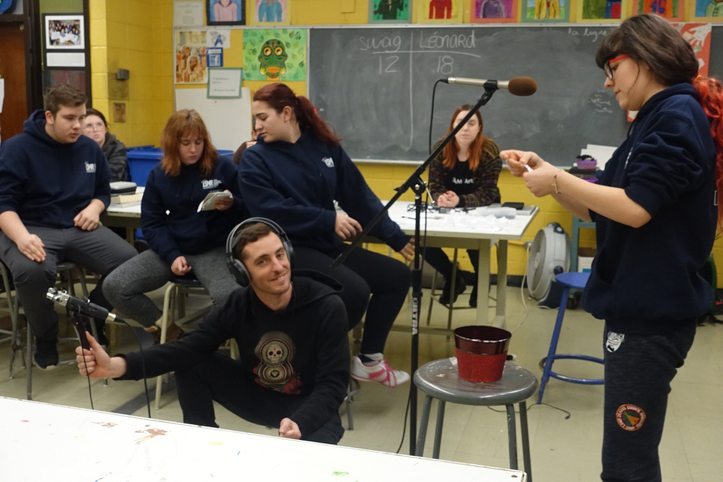 Jeunes adolescents dans une salle de classe en train de faire un projet ou une présentation, certains assis ou debout, avec matériel de classe autour.