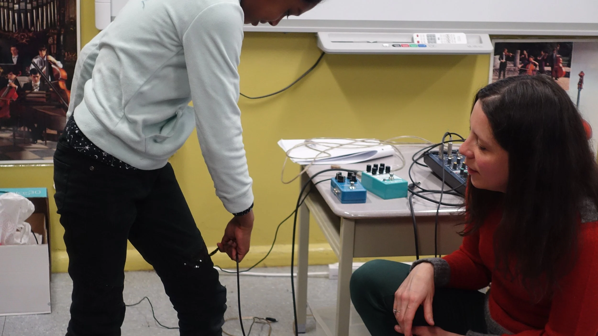 Une jeune personne en sweatshirt gris manipule un câble électrique, tandis qu'une femme aux cheveux longs, portant un pull rouge, regarde attentivement. La scène se déroule dans une pièce avec une table portant des équipements électroniques, notammen