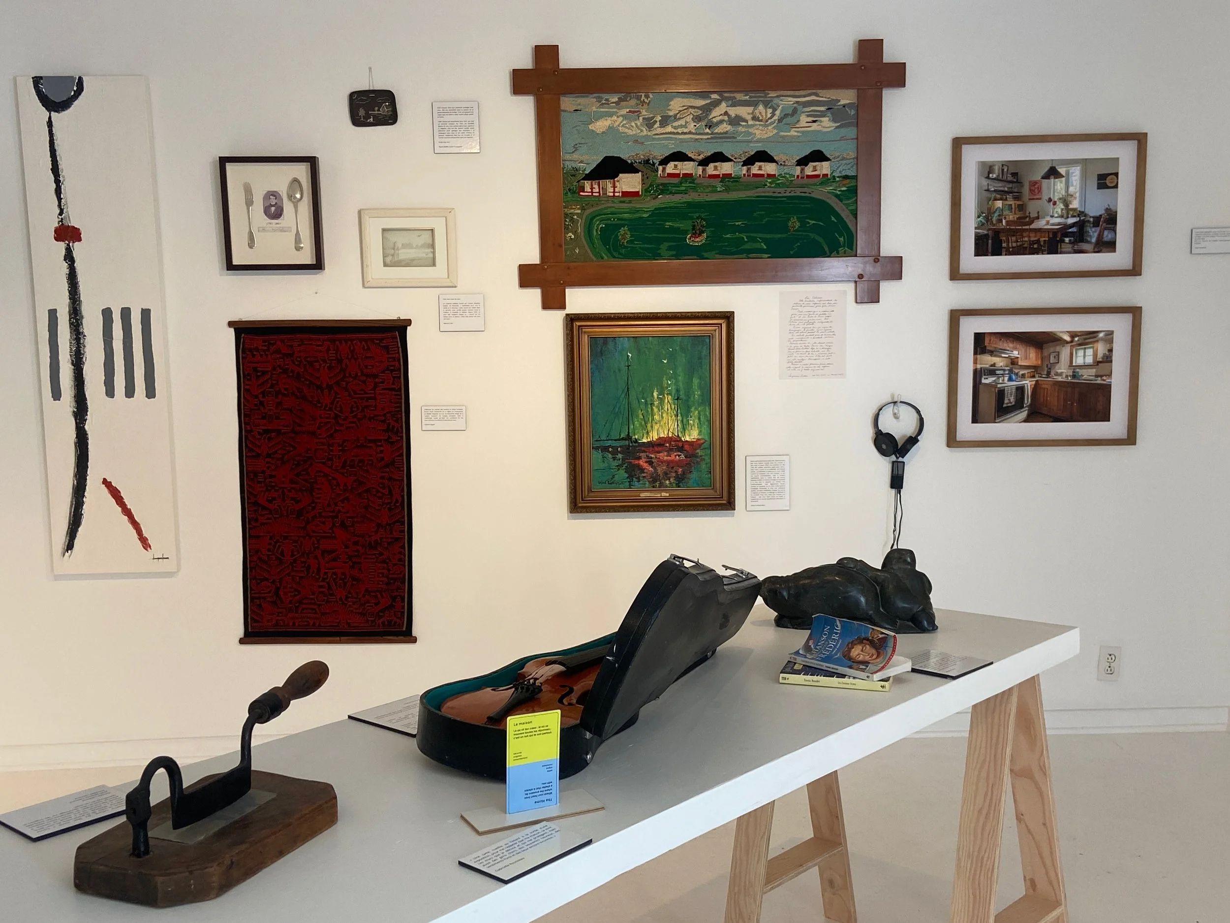 Une exposition d'art avec plusieurs œuvres murales, peintures, un tapis rouge, un casque audio suspendu, et un bureau avec un violon dans une valise et quelques livres.