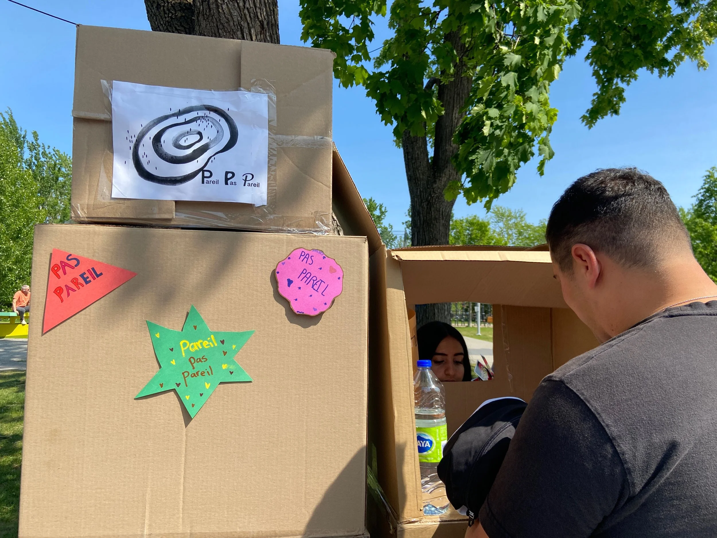 Un stand en carton avec des panneaux colorés indiquant "Pas pareil", où une jeune femme regarde à travers une petite fenêtre, avec un homme en face, sous un arbre en plein air par temps ensoleillé.