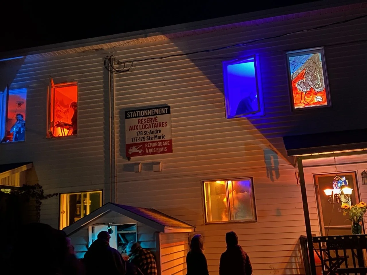 Une maison éclairée avec des fenêtres colorées une à une bleu, une rouge, une orange et une autre jaune, à la nuit. Des personnes sont assises ou debout à l'extérieur de la maison dans l'obscurité.