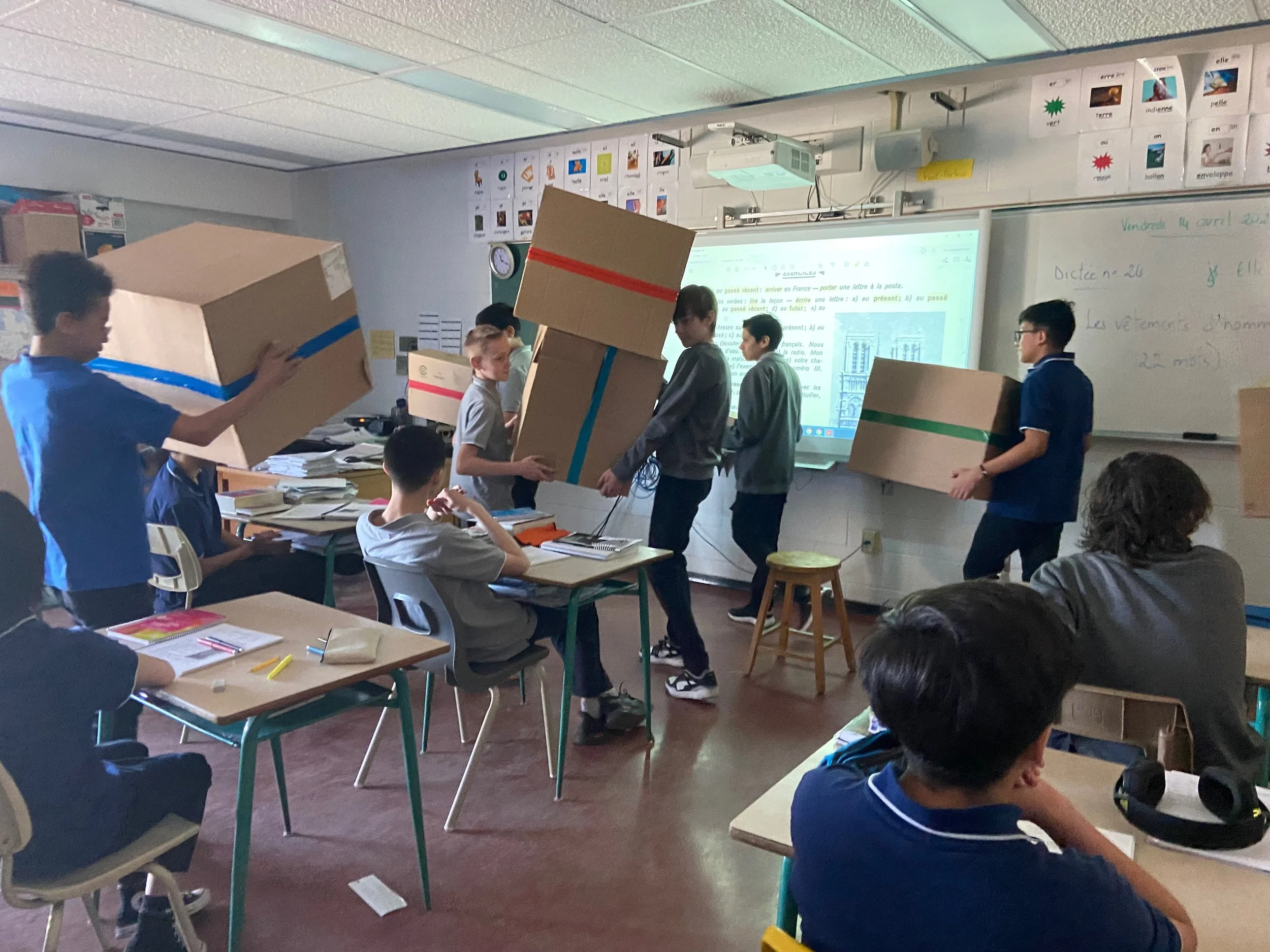 Des élèves de classe utilisant de grandes boîtes en carton dans une salle de classe, certains tenant les boîtes, d'autres assis ou se tenant debout, avec un tableau blanc et un projecteur à l'avant.
