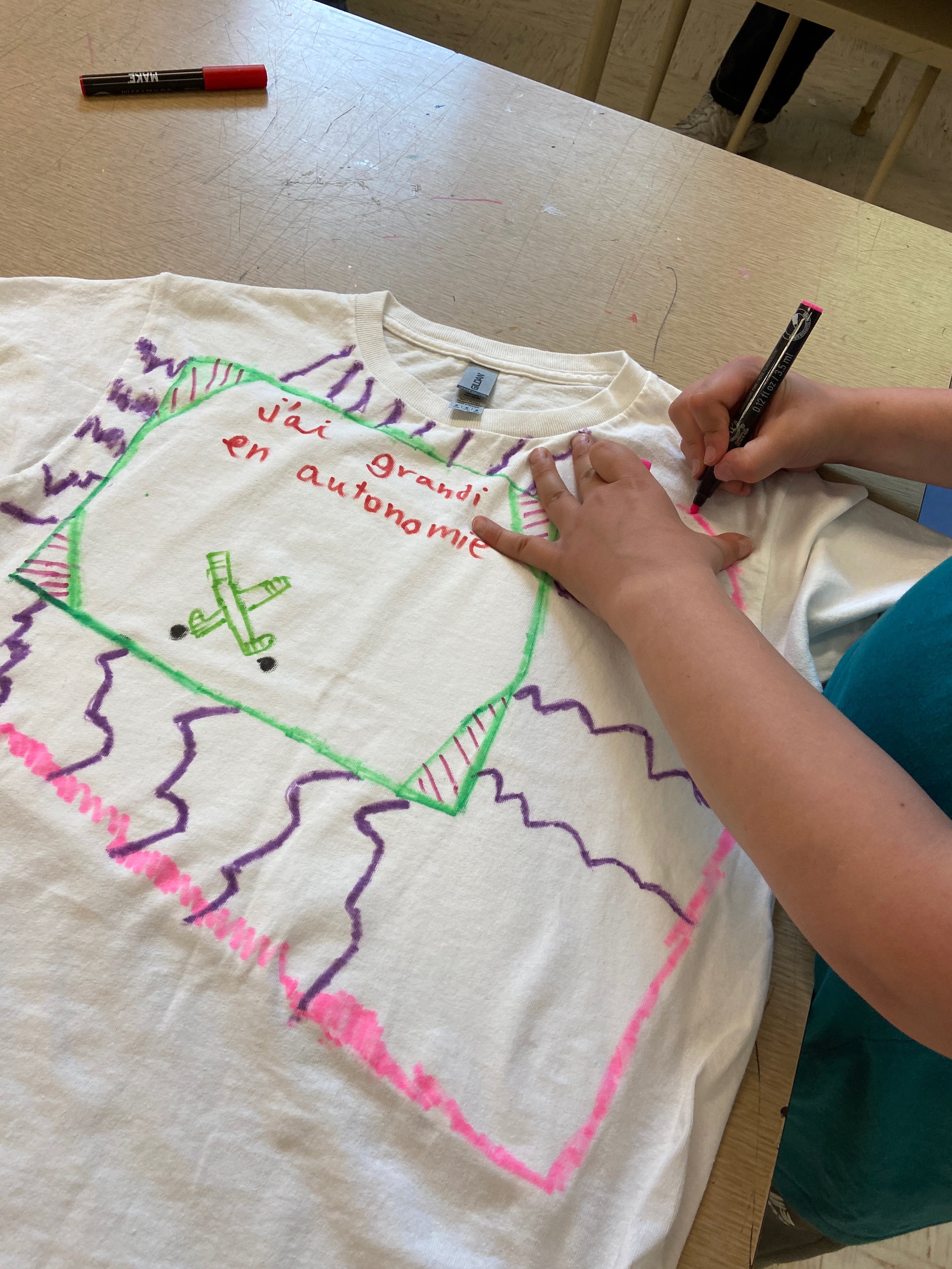 Une personne écrit sur un t-shirt blanc avec un marqueur noir, des dessins colorés et le texte 'j'ai grandi en autonomie'.