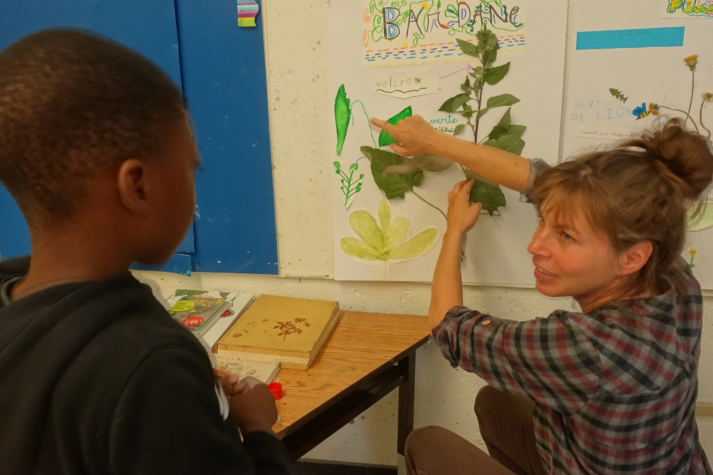 Une femme explique à un jeune garçon une plantes dans une salle de classe. La femme montre une plante ressemblant à une feuille de basilic ou de menthe, placée contre un tableau avec des dessins et des écrits."BARDINE" est écrit en couleur en haut. L