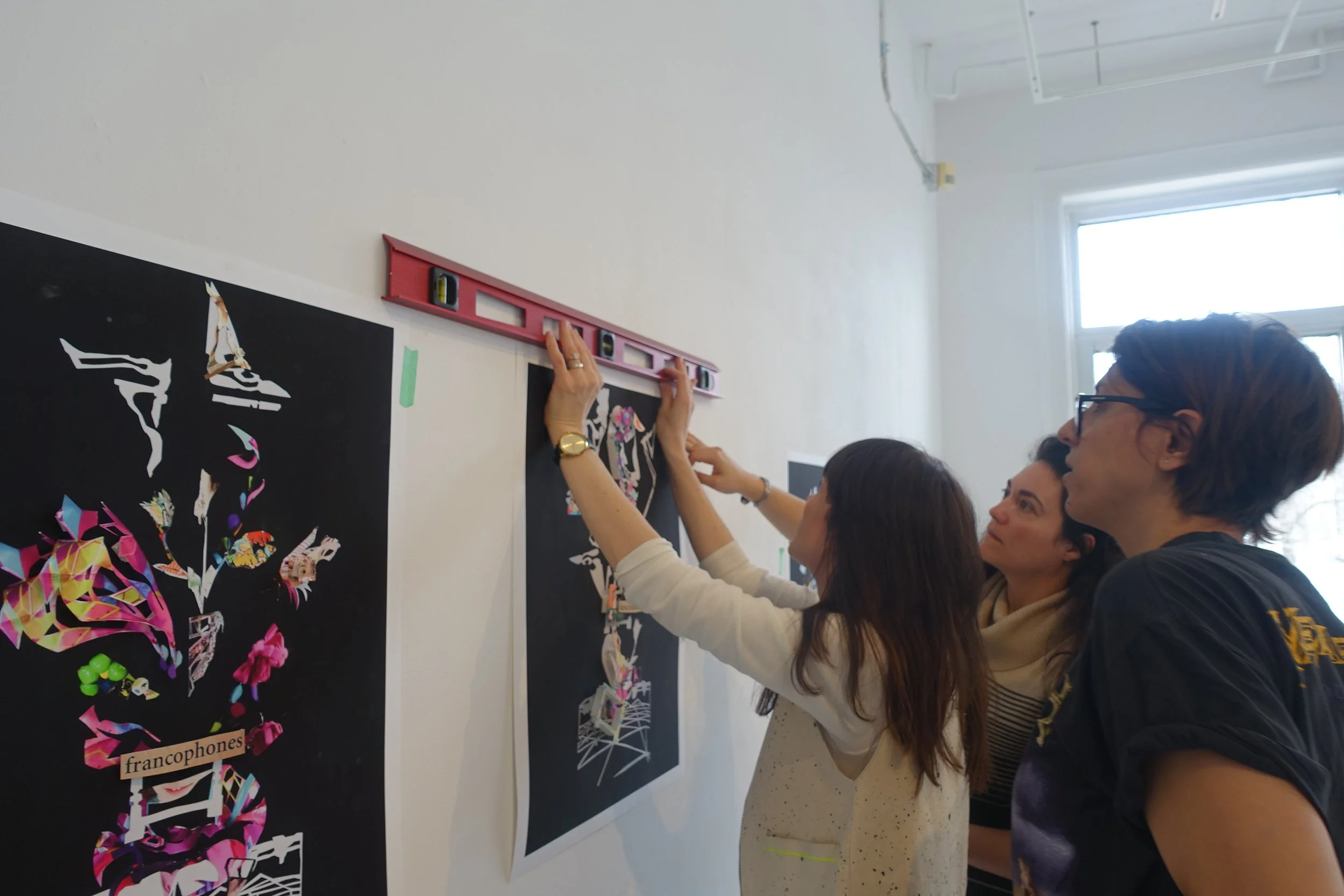 Trois personnes accrochant une œuvre d'art sur un mur blanc à l'aide d'un niveau laser. D'autres œuvres d'art abstraites sont affichées sur le mur.