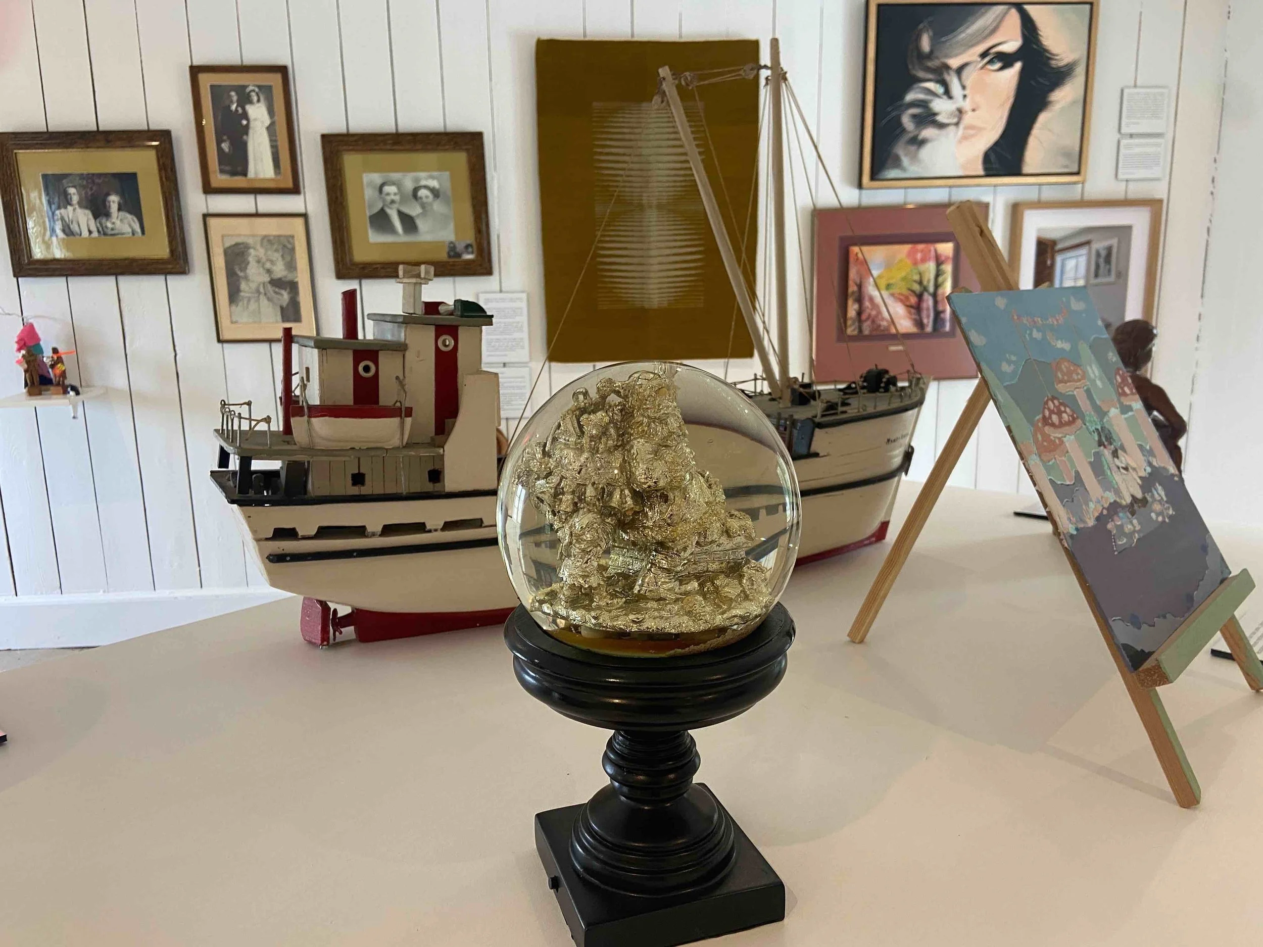 Une exposition avec des modèles de bateaux, une boule en verre contenant un objet en or, des tableaux sur le mur et une sculpture d'une femme avec un chat.
