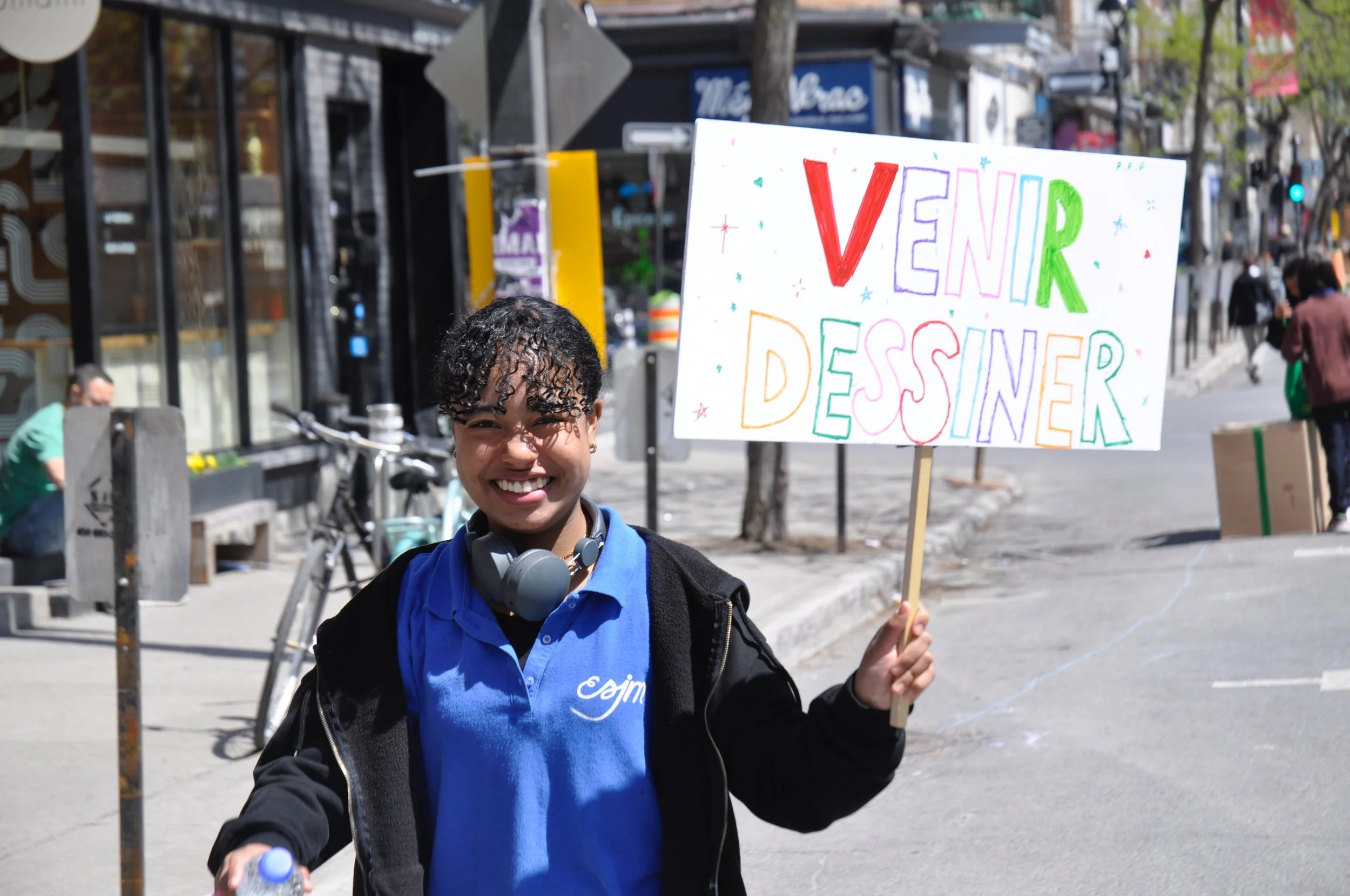 Jeune femme souriante tenant une pancarte colorée qui dit "VENEIR DESSINER", marché urbain avec des magasins et des piétons en arrière-plan.