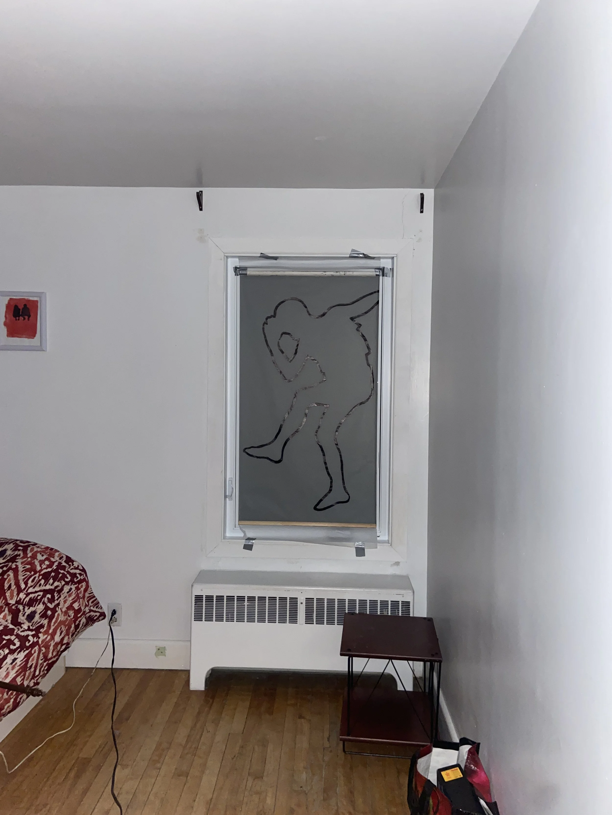 Une pièce avec un mur blanc, un radiateur en dessous d'une fenêtre. Sur la fenêtre, il y a un dessin noir d'une figure humaine sculptée ou dessinée, ressemblant à une silhouette déhanchée. À gauche, un cadre rouge est accroché au mur. Un meuble en bois avec une structure métallique se trouve à droite. Au sol en bois clair, une prise électrique avec un câble suspendu est visible, ainsi qu'un sac ou une boîte en bas à droite.