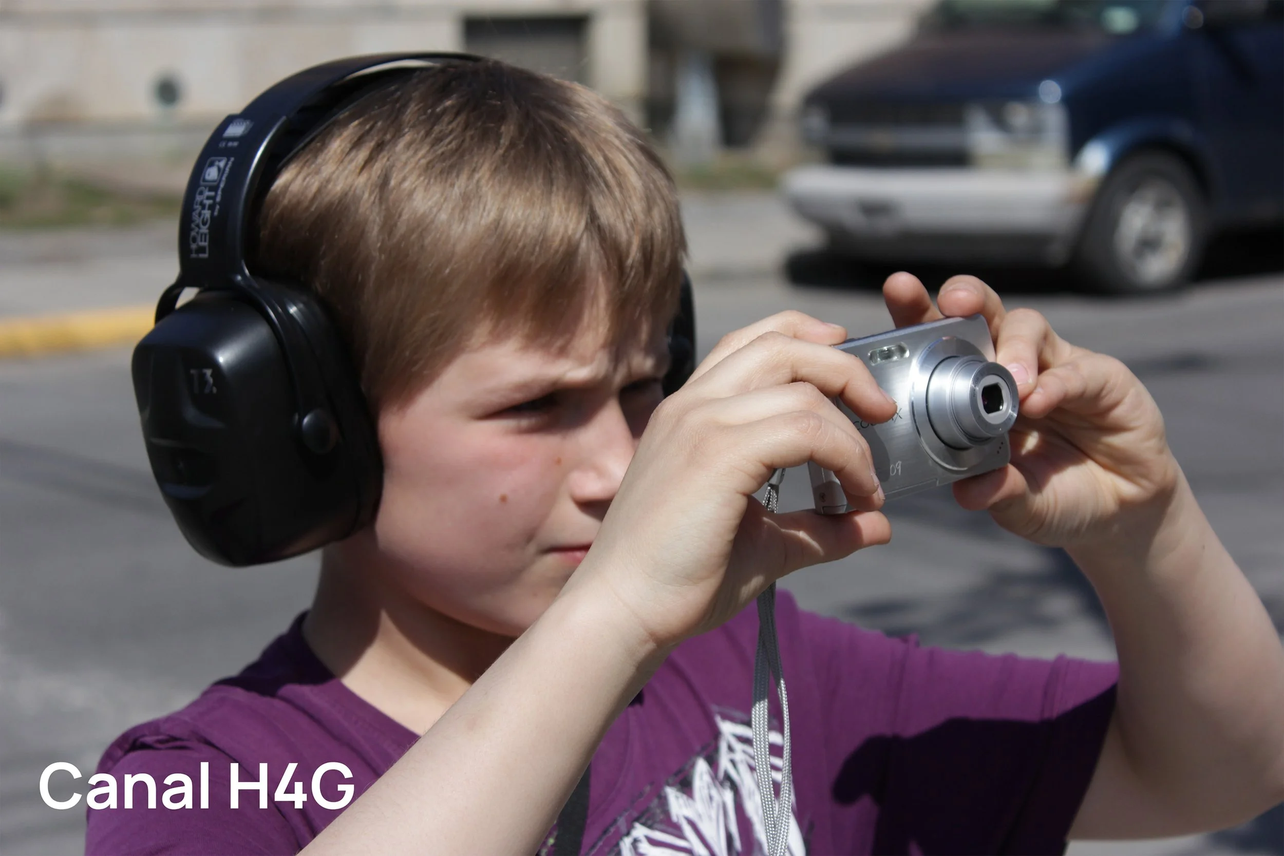 Un garçon portant des écouteurs noirs et tenant un appareil photo argenté, en train de prendre une photo dans une rue ensoleillée, avec des voitures en arrière-plan.