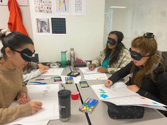 Quatre personnes assises à une table d'école portant des masques oculaires colorés, travaillant sur des projets avec du papier, des crayons et des fournitures artisanales dans une salle de classe.