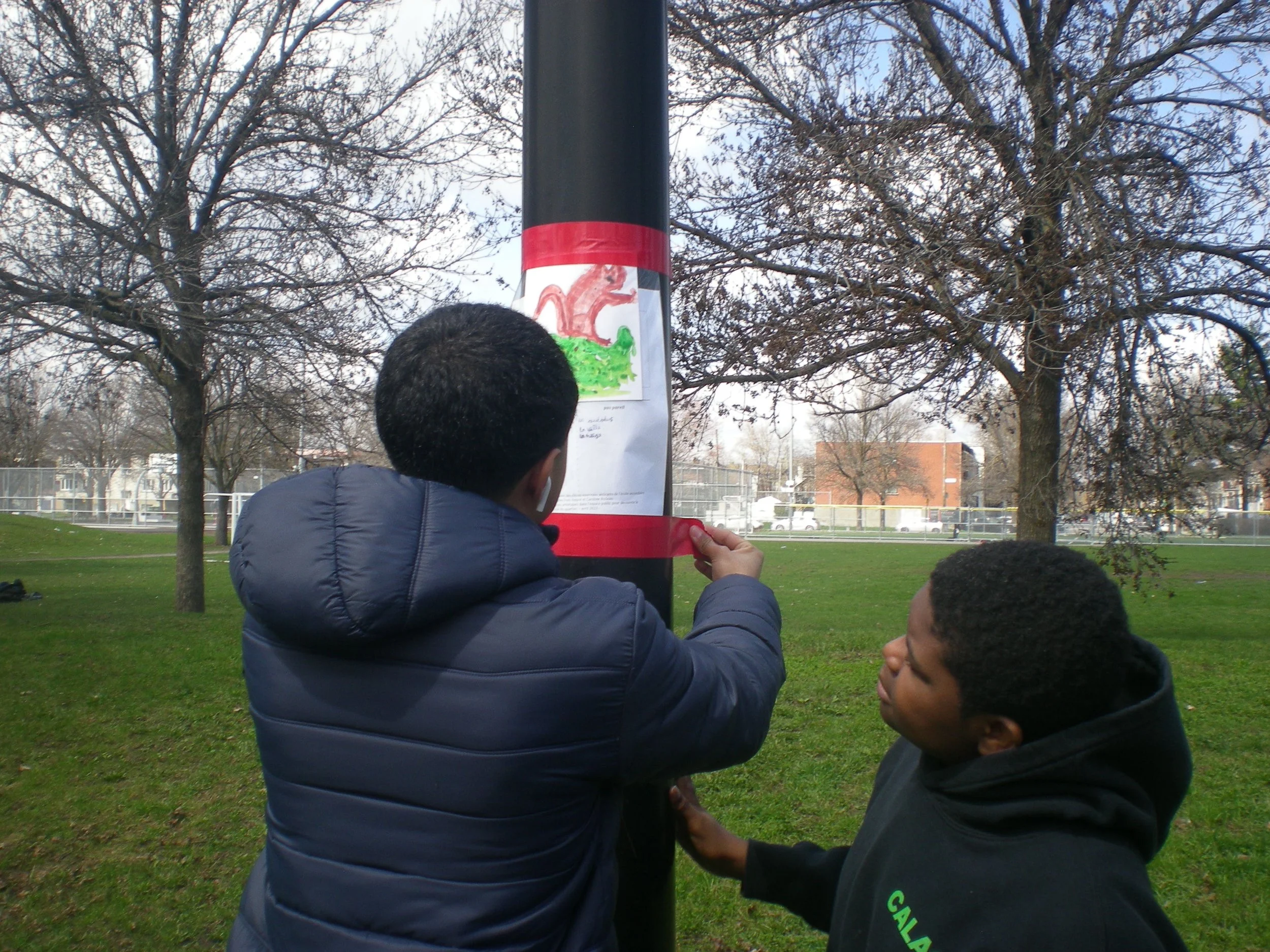 Deux garçons collent une affiche colorée sur un poteau dans un parc, avec des arbres nus en arrière-plan.