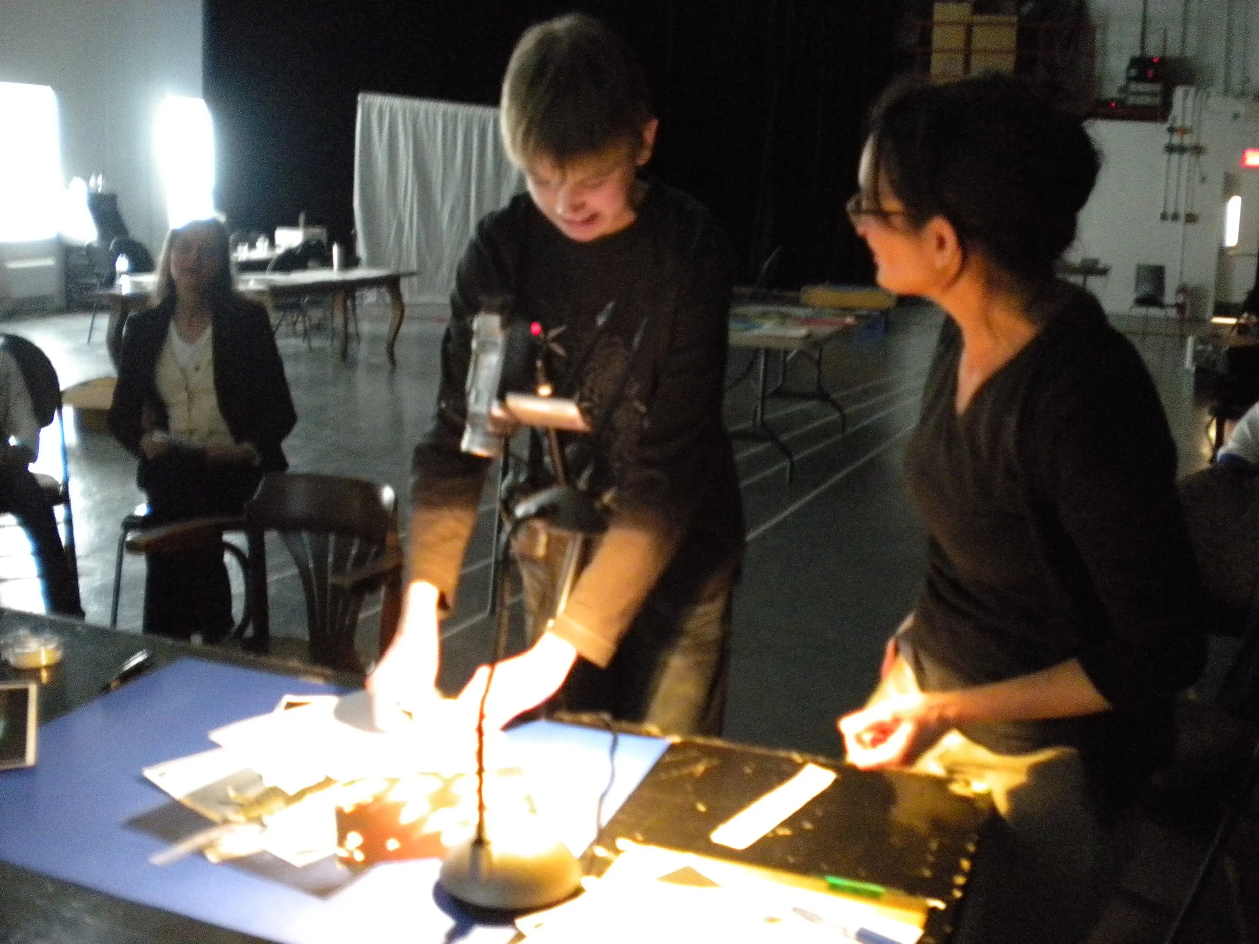 Un jeune garçon et une femme en gilet noir parlent et manipulent un robot lumineux sur une table dans une pièce sombre. Une femme en arrière-plan assise observe.