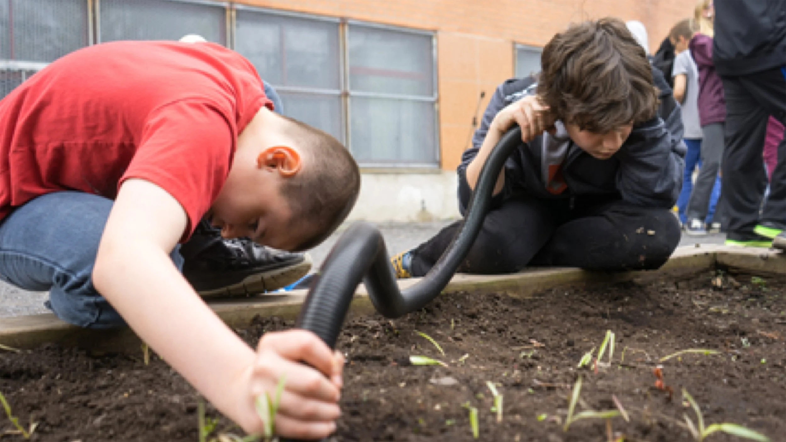 Deux enfants plantent des jeunes pousses dans la terre dans une allée de jardin, l'un à terre utilisant un outil pour creuser la terre, l'autre à genoux tenant un tuyau d'arrosage, en extérieur, avec d'autres enfants en arrière-plan.