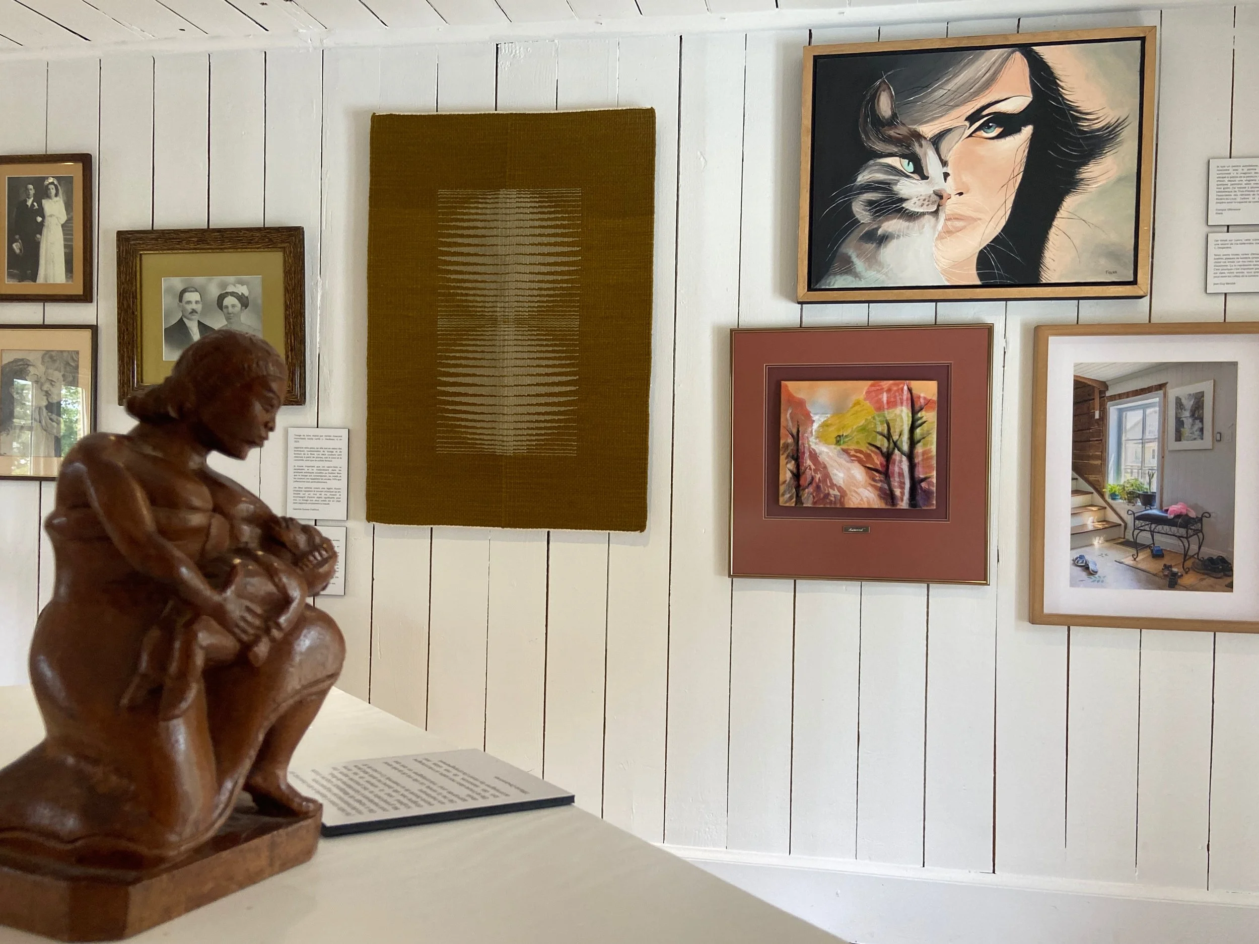 Exposition d'art avec plusieurs tableaux exposés sur un mur blanc, notamment un portrait stylisé d'une femme avec un chat, une peinture de paysage avec des arbres, et une photo de scène intérieure d'une entrée de maison. Sculpture en bois d'une femme