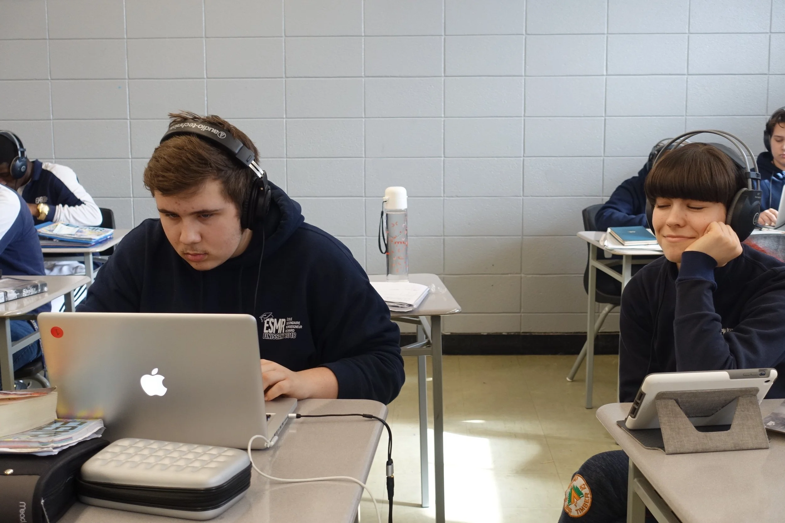 Deux adolescents assis à des tables d'école, portant des écouteurs et utilisant des appareils électroniques dans une salle de classe.