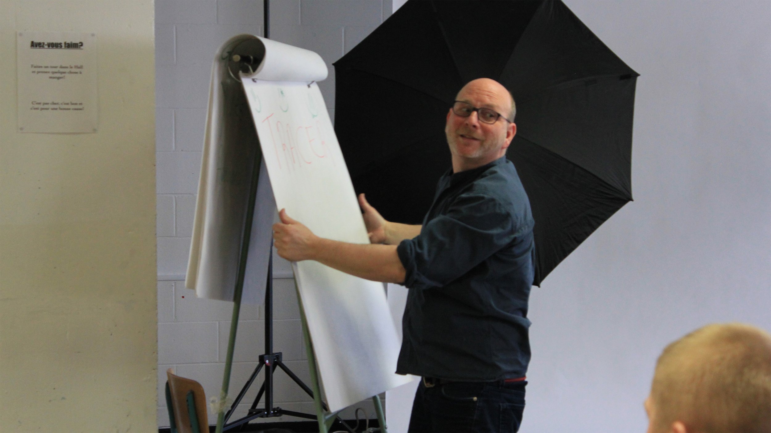 Un homme souriant portant des lunettes, qui écrit sur un tableau blanc près d'un grand parapluie noir dans une salle