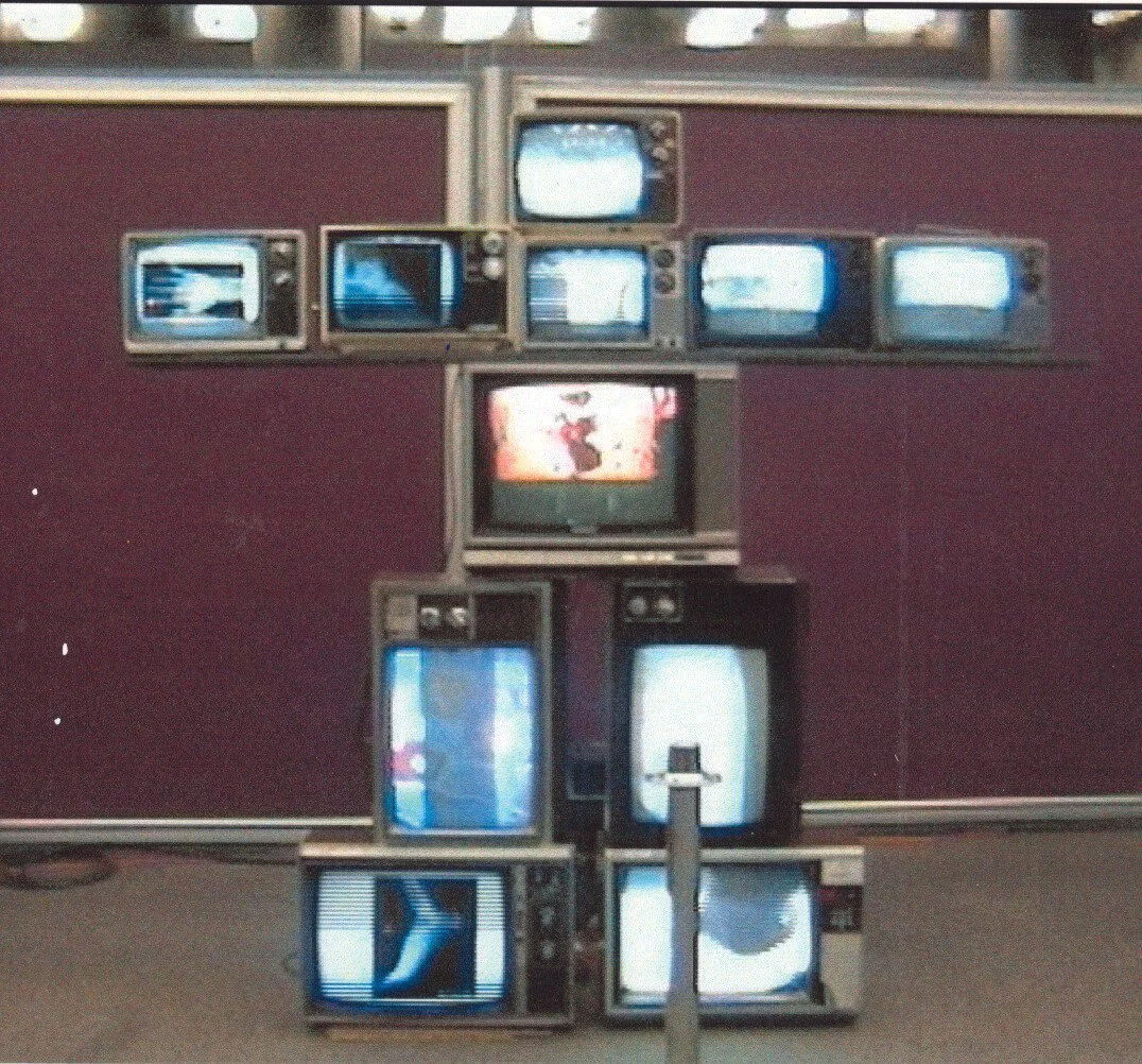 Une sculpture composée de plusieurs vieux téléviseurs empilés formant une croix sur un mur violet.