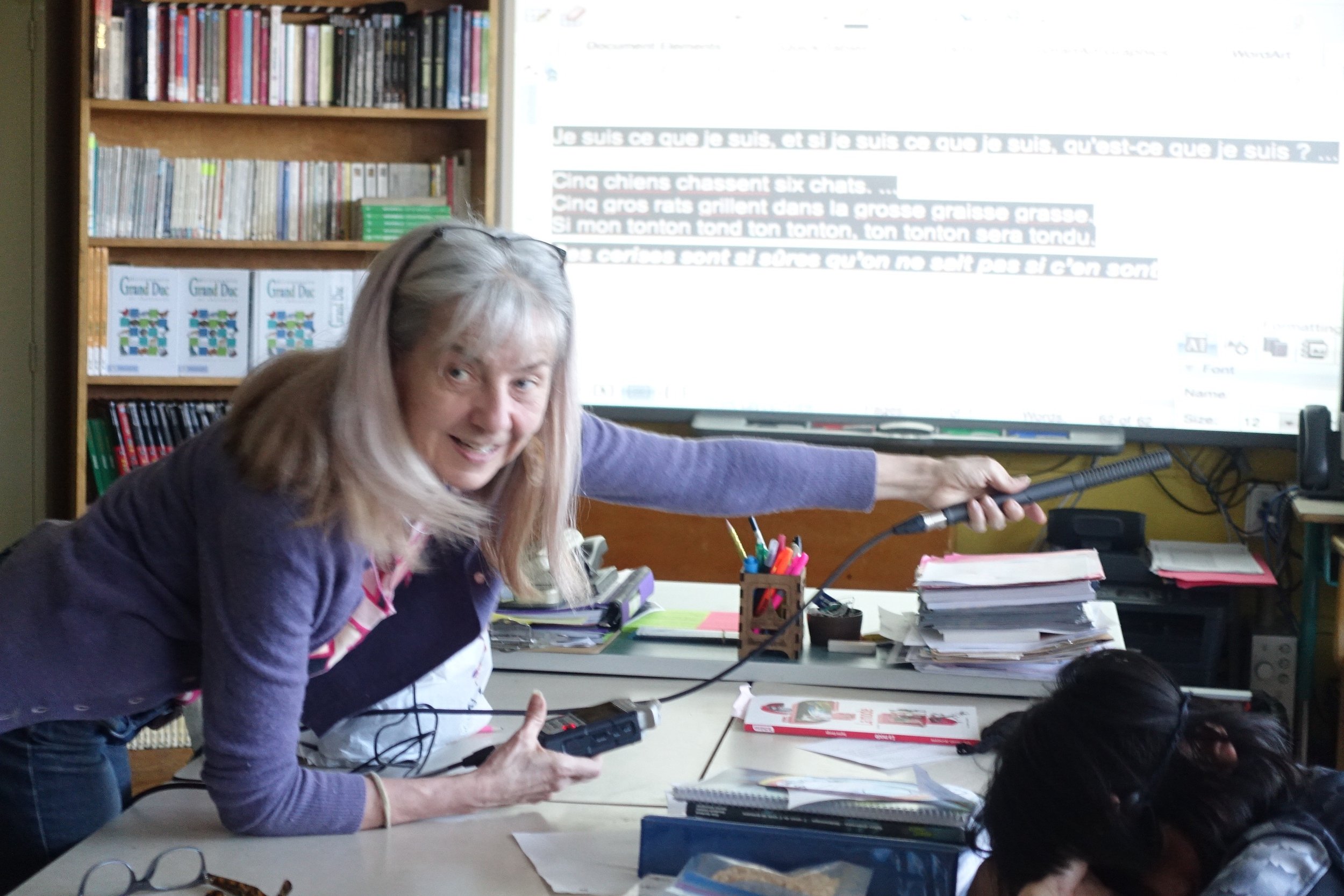 Une femme souriante pointant un micro vers une personne assise à une table encombrée de livres et de papiers, dans une salle de classe ou bibliothèque avec étagères pleines de livres en arrière-plan.