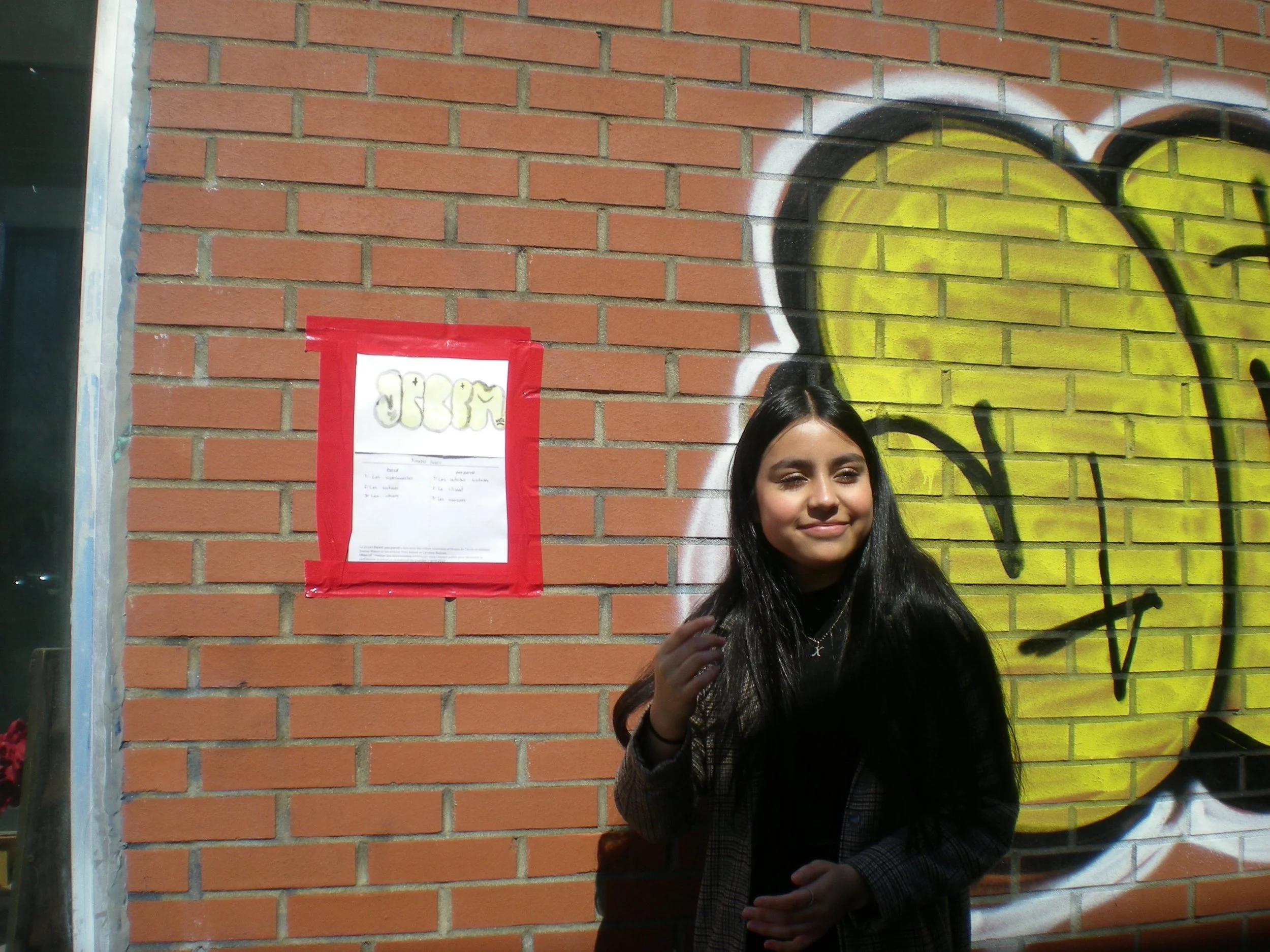 Une jeune femme souriante avec des cheveux longs noirs devant un mur en briques rouge avec un graffiti jaune et noir et une affiche encadrée en rouge.