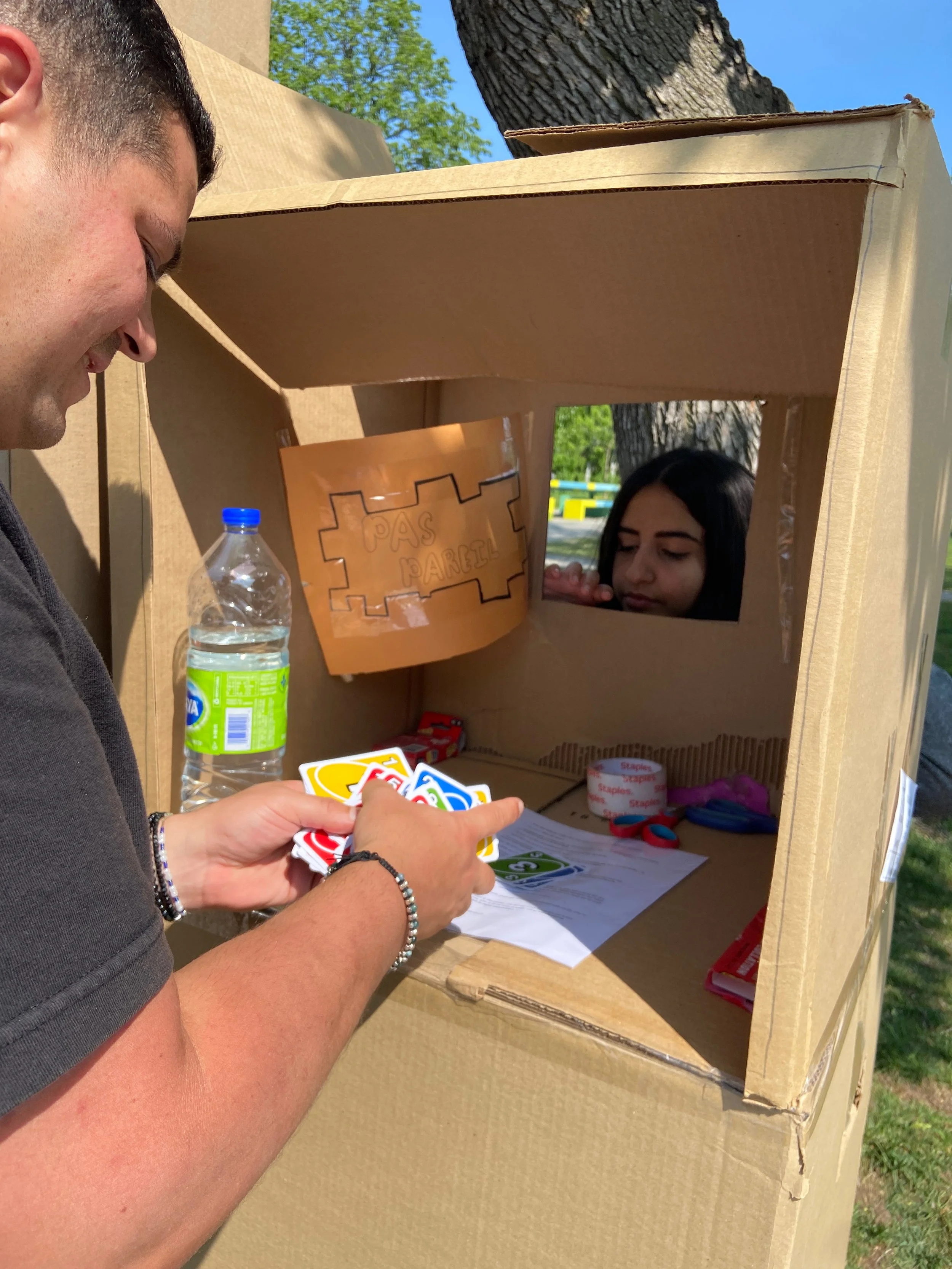 Une personne joue à un jeu de cartes dans une boîte en carton transformée en stand de jeu, avec une fille regardant par une fenêtre découpée, à l'extérieur par une journée ensoleillée.