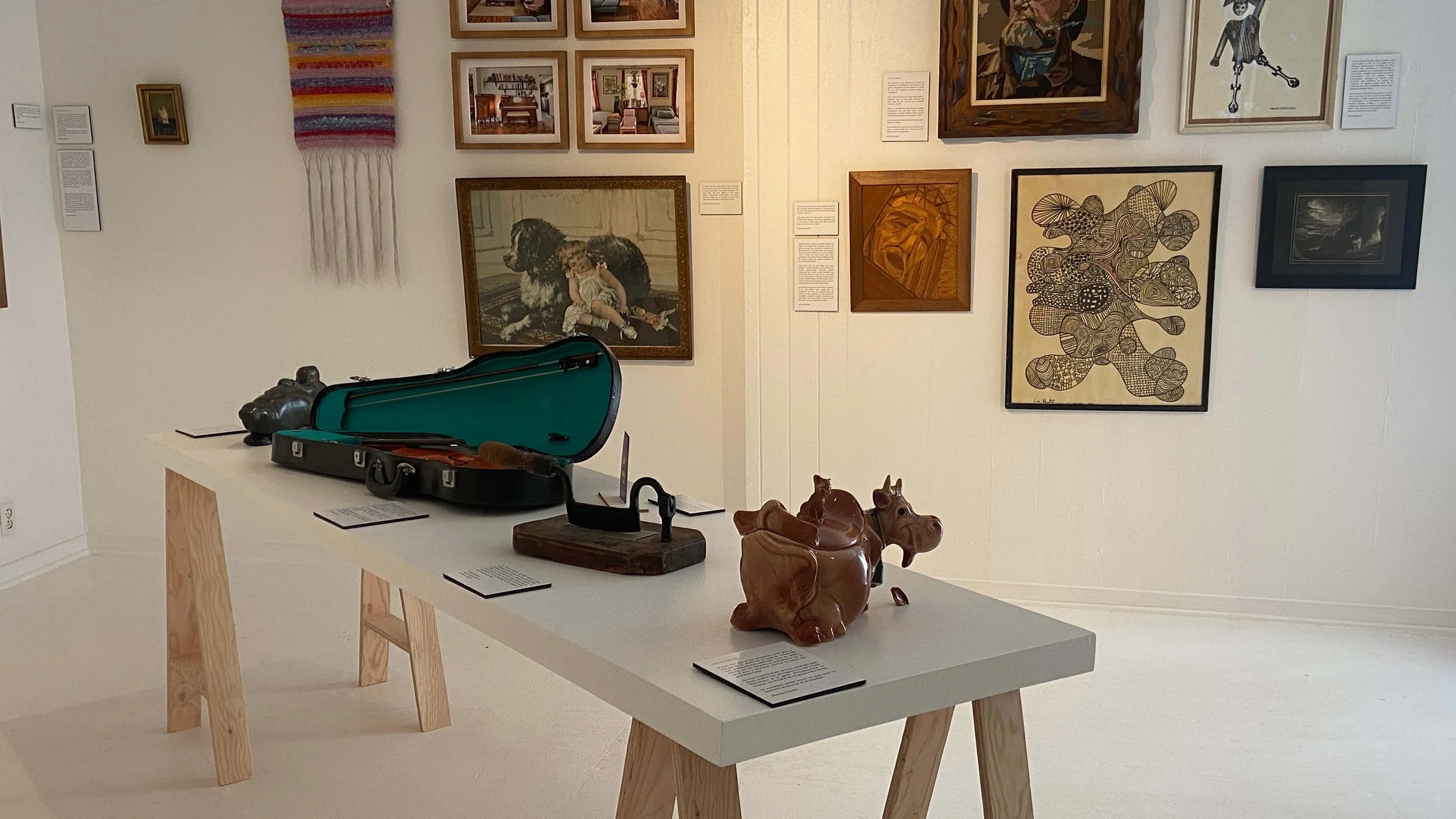 Exposition d'objets artistiques et de peintures dans une galerie, comprenant un sac de violon, des sculptures en bois, un violon dans une boîte, et plusieurs tableaux sur le mur.