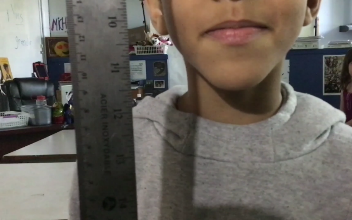 Jeune garçon portant un sweat gris, tenant une règle de 30 cm, dans une salle de classe avec des meubles et des posters à l'arrière-plan.