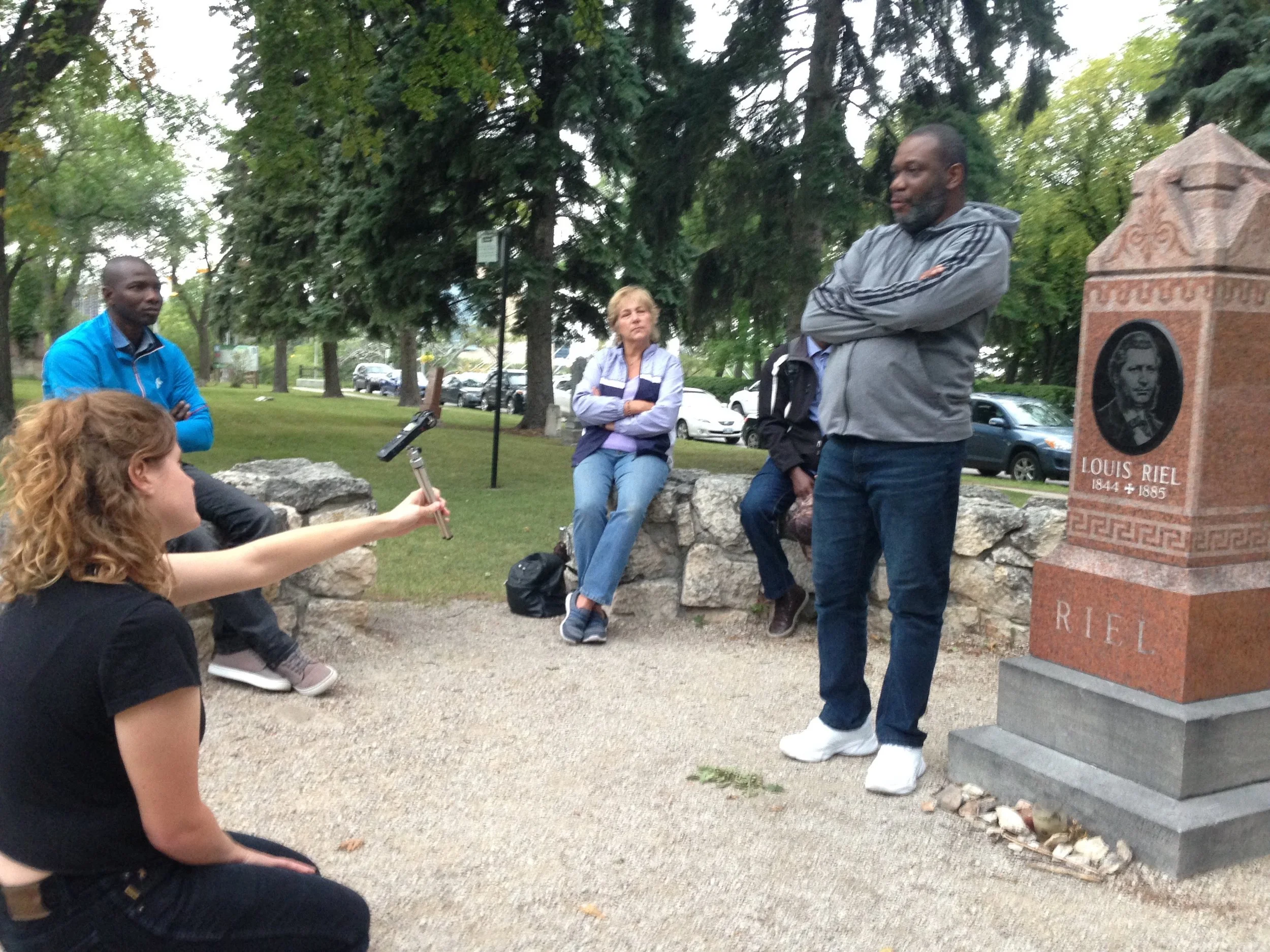 Un groupe de personnes, dont un homme noir et une femme blanche, écoute une femme aux cheveux roux prendre un selfie avec un bâton de selfie. La scène se déroule dans un parc avec des arbres et une pierre tombale en arrière-plan, portant le nom 'Loui