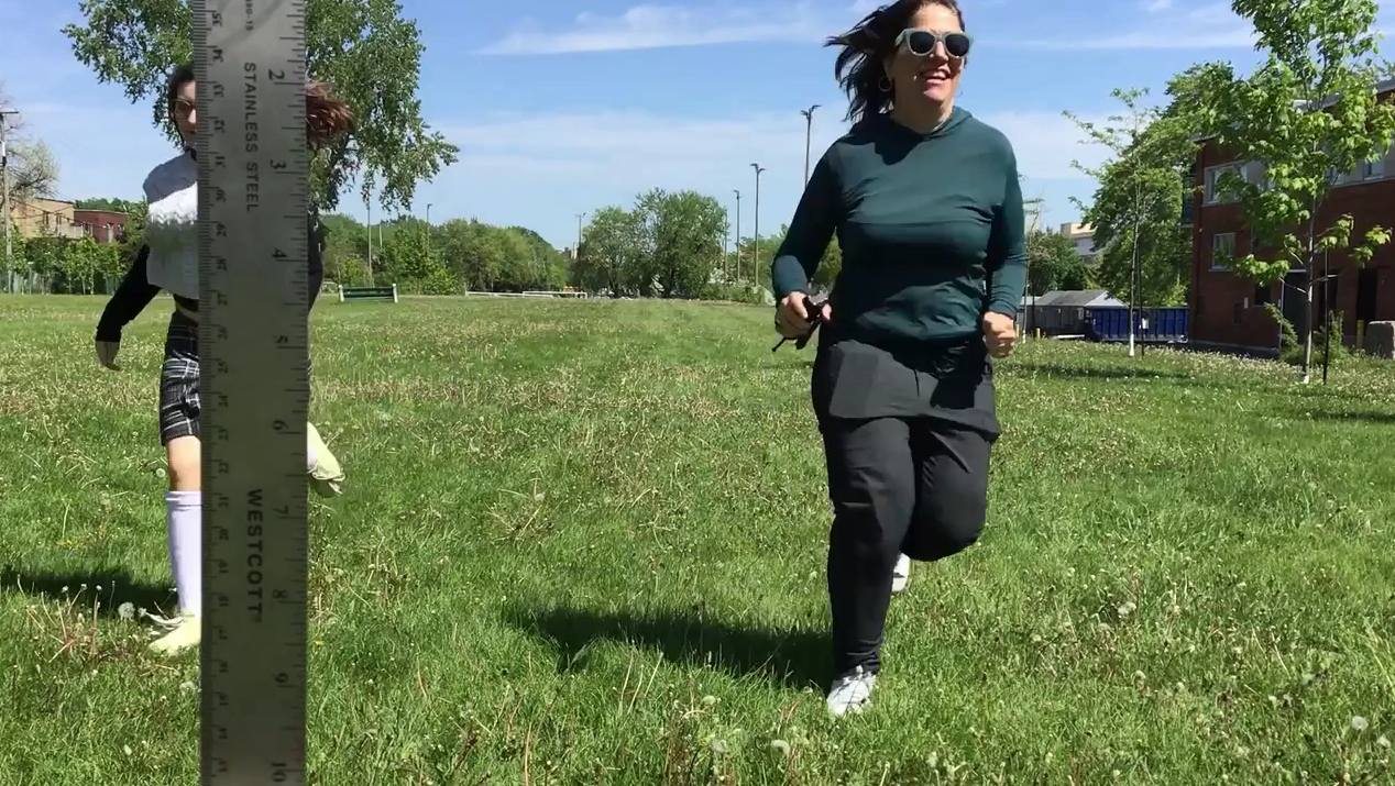 Deux femmes courent dans un parc vert ensoleillé, portant des vêtements de sport et des lunettes de soleil, avec des bâtiments et des arbres en arrière-plan.