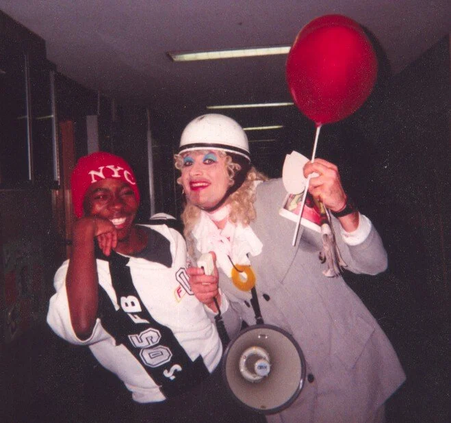 Deux personnes déguisées en clowns, souriantes, dans un décor intérieur, l'une avec un ballon rouge.