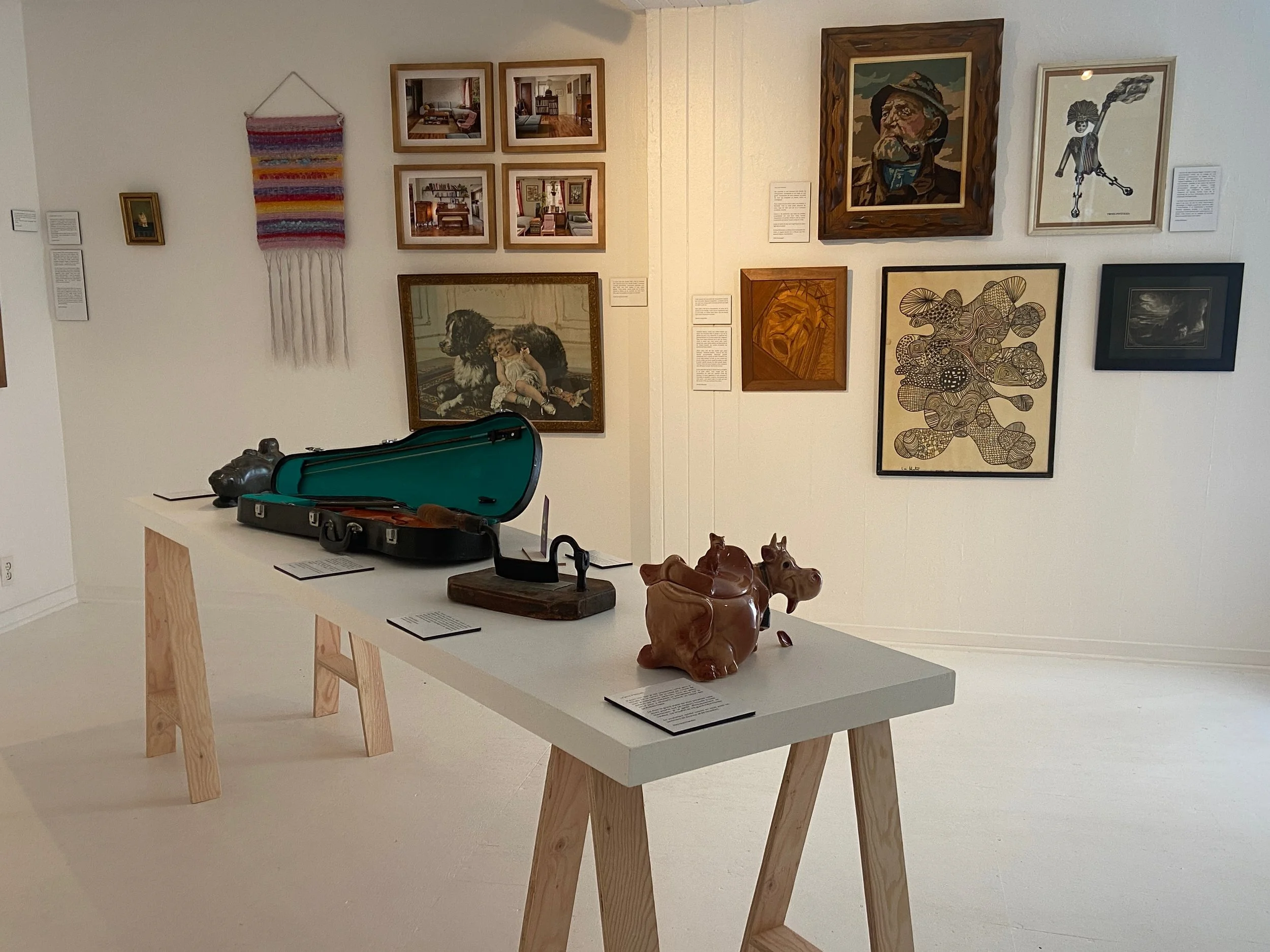 Exposition d'œuvres d'art, comprenant des tableaux, une sculpture en céramique de forme animale, un étui à violon ouvert avec un violon à l'intérieur, et un tableau en tissus colorés suspendu, dans une galerie d'art.