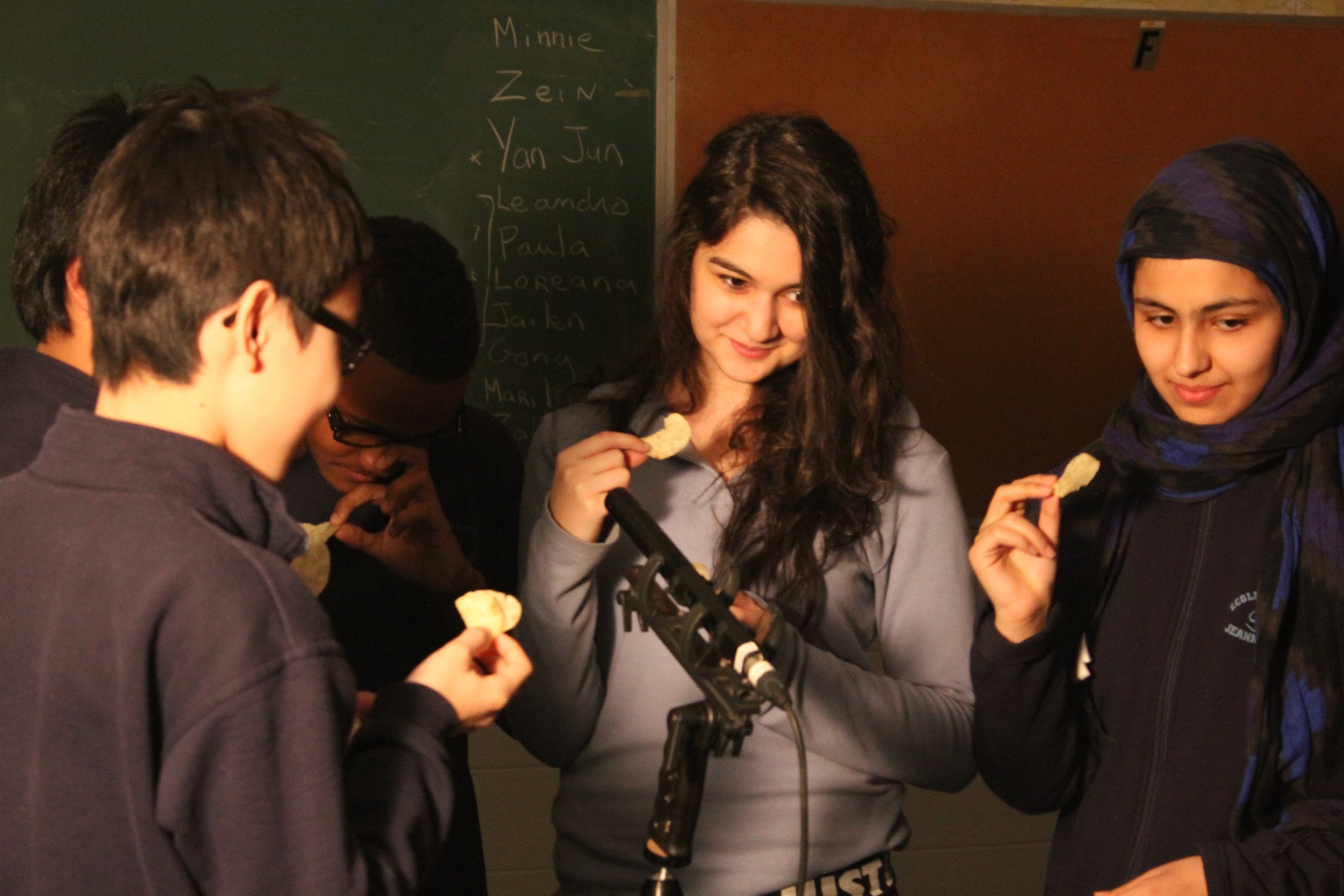 Groupe de jeunes filles et garçons, certains portant des lunettes et foulards, dégustant des chips dans une salle avec tableau noir en arrière-plan.